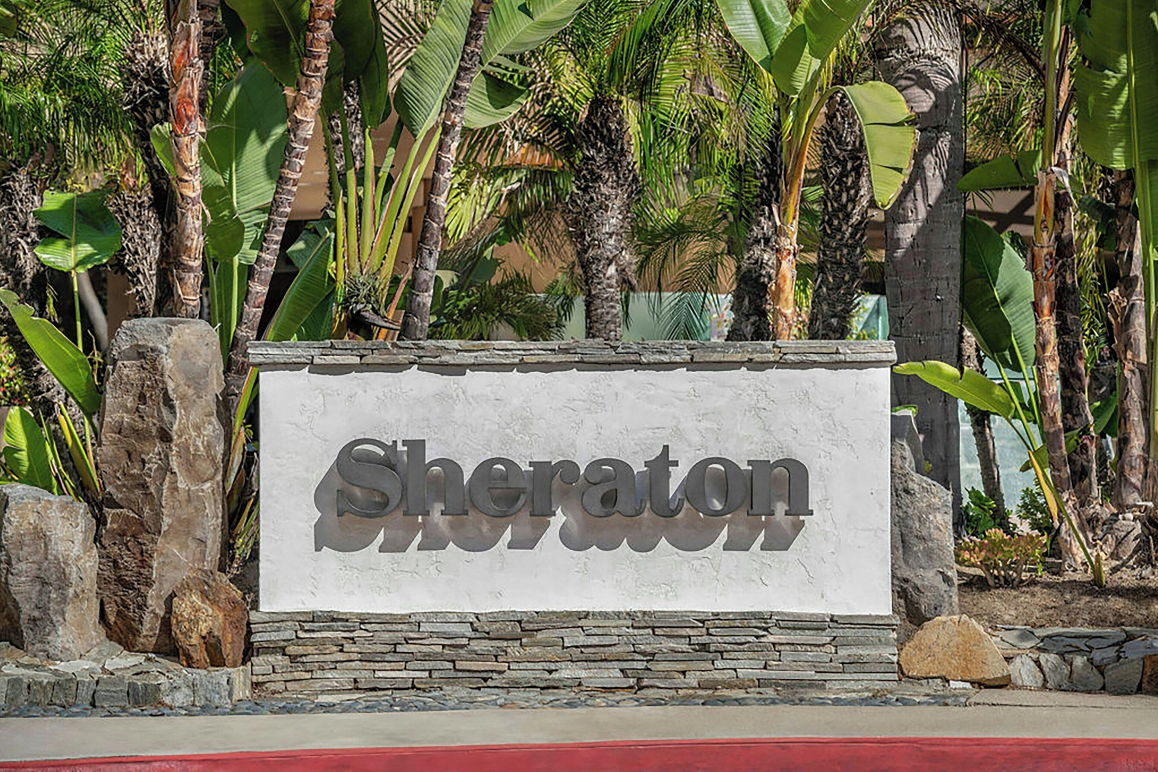 A sign with the word "Sheraton" in a bold, sans-serif font, surrounded by tropical plants and rocks.
