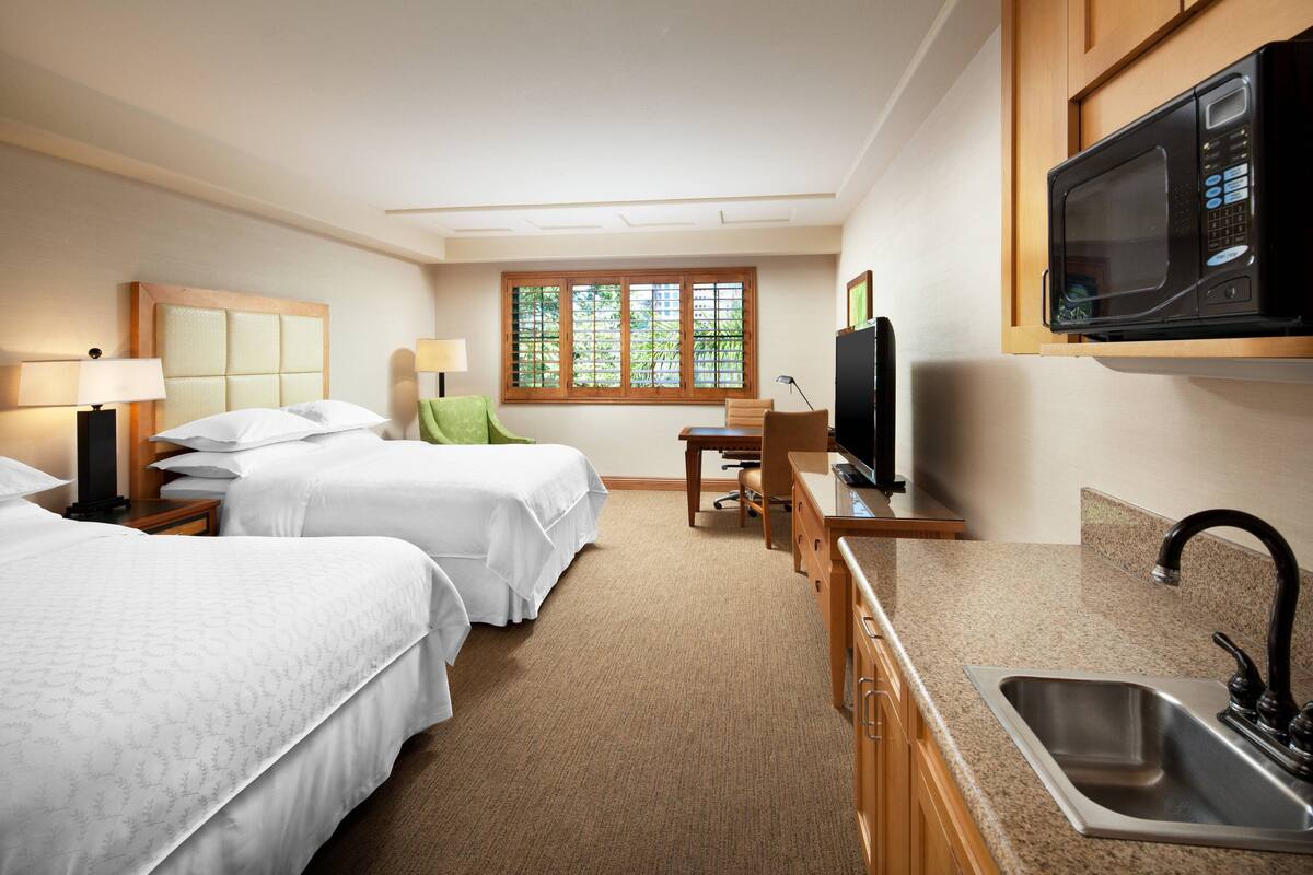 A hotel room with two beds, a desk with a chair, a television, a microwave, and a sink.