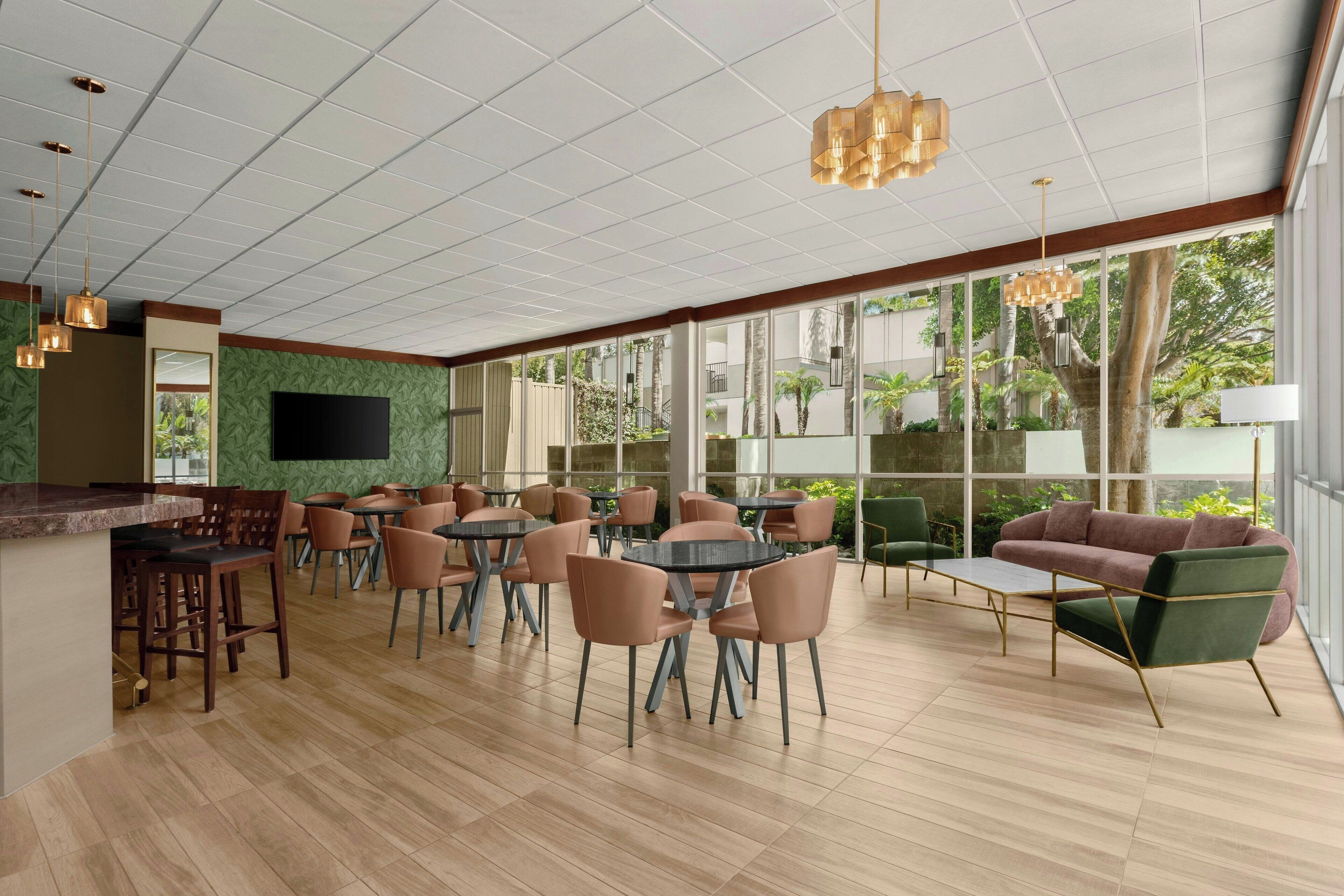 A spacious indoor area with a bar, multiple tables and chairs, and large windows offering a view of greenery outside.
