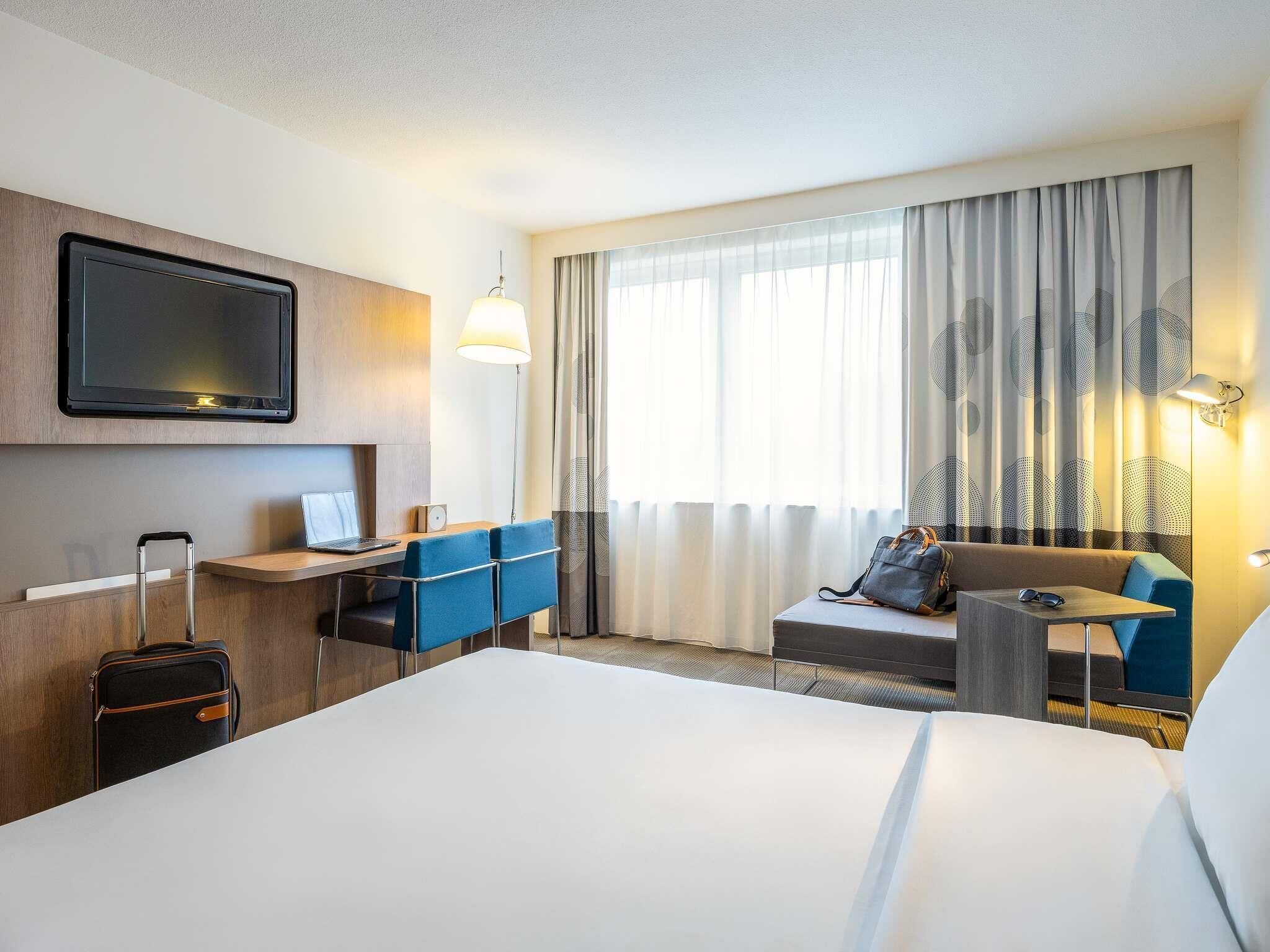 Une chambre d’hôtel avec un lit, un bureau, des chaises, une télévision, une valise et une fenêtre avec des rideaux.