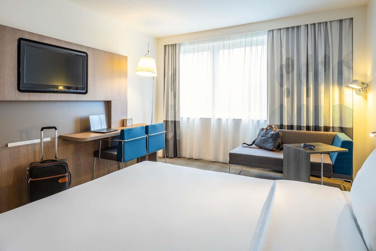 A hotel room with a bed, desk, chairs, a TV, a suitcase, and a window with curtains.