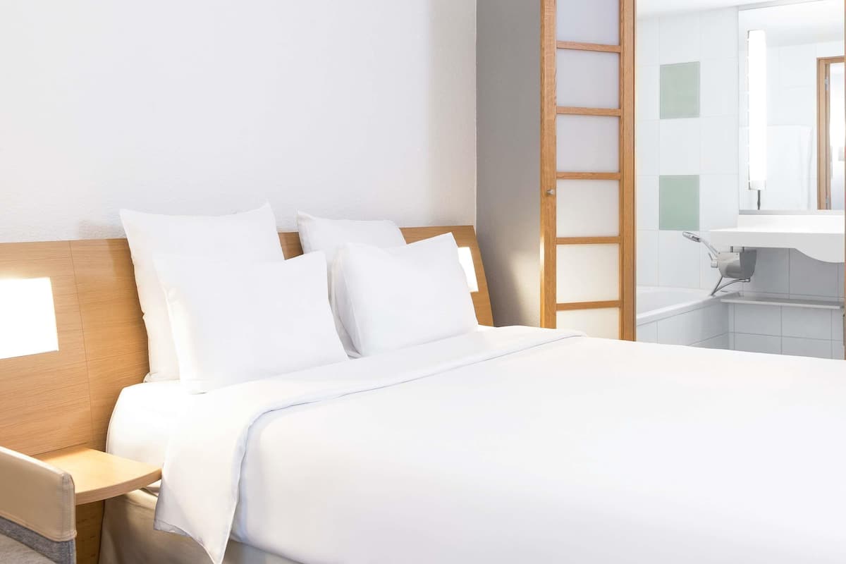 A hotel room with a bed, a wooden headboard, a nightstand, a bathroom with a sink and mirror, and a sliding door.