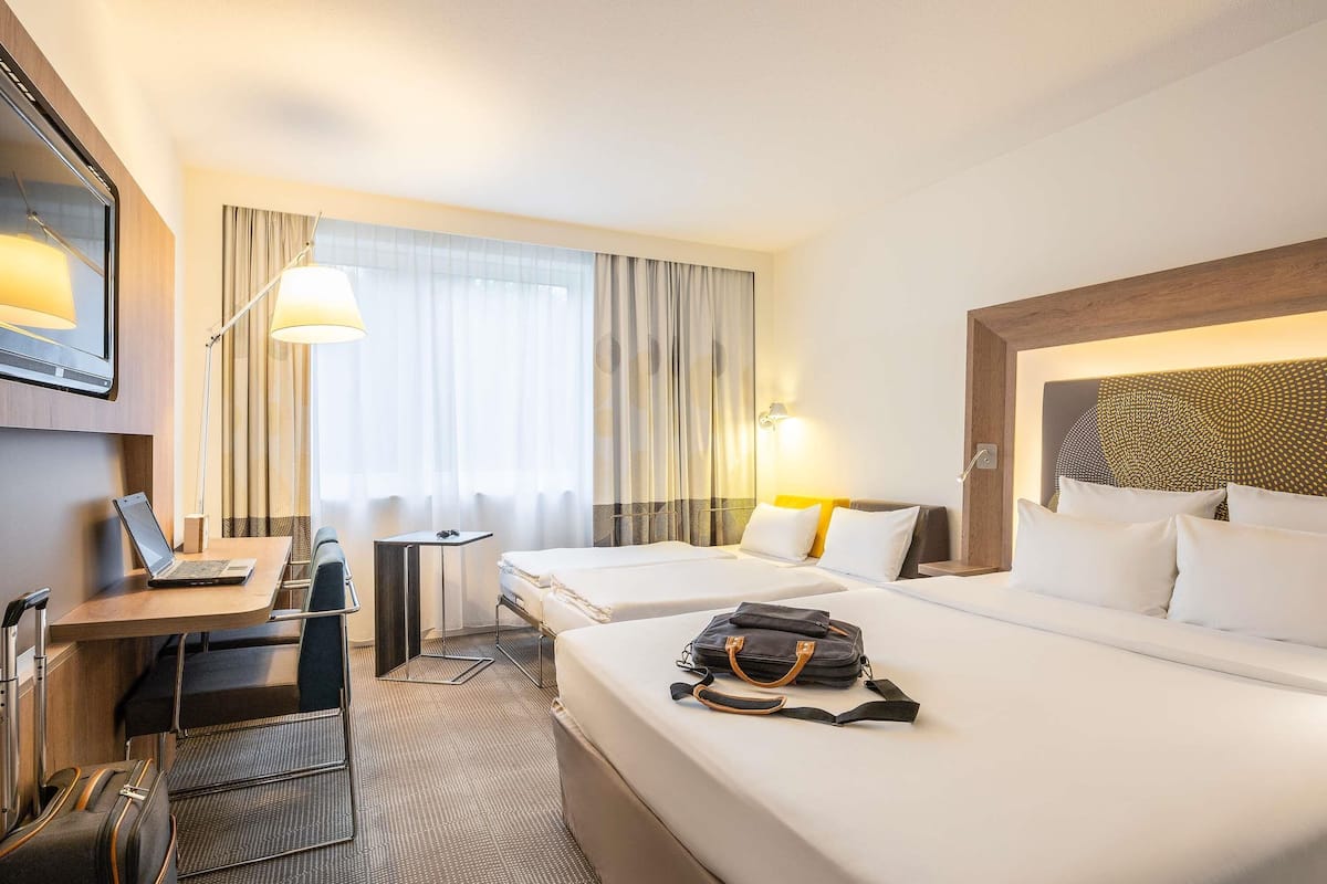 A hotel room with two beds, a desk with a laptop, a chair, a suitcase, and a window with curtains.
