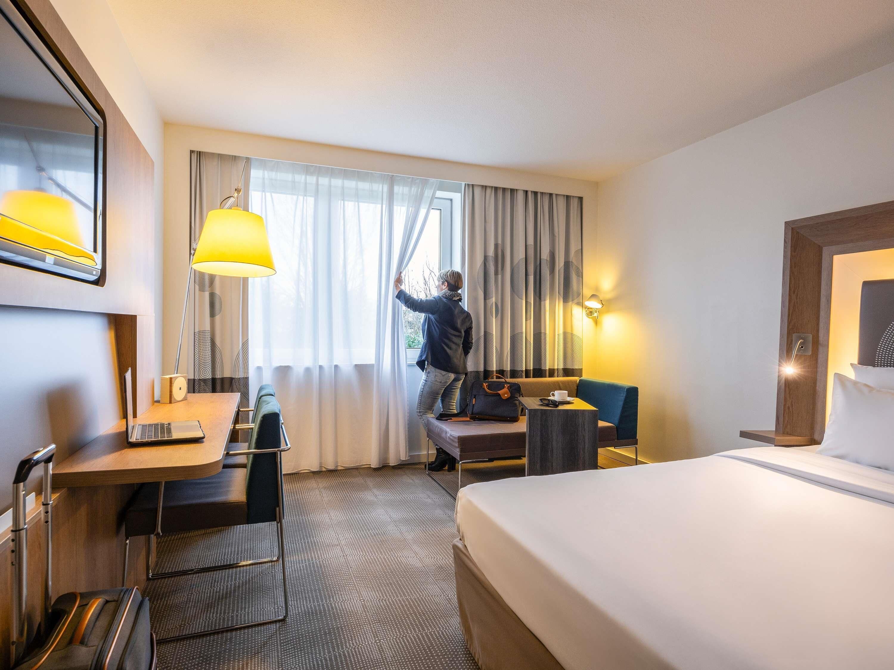 A hotel room with a bed, a desk with a laptop, a chair, a suitcase, and a person adjusting curtains.