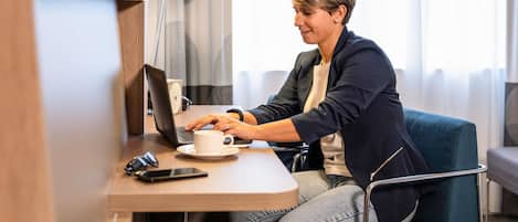 A person using a laptop at a desk in a hotel room with a lamp, a cup, and a smartphone on the table.