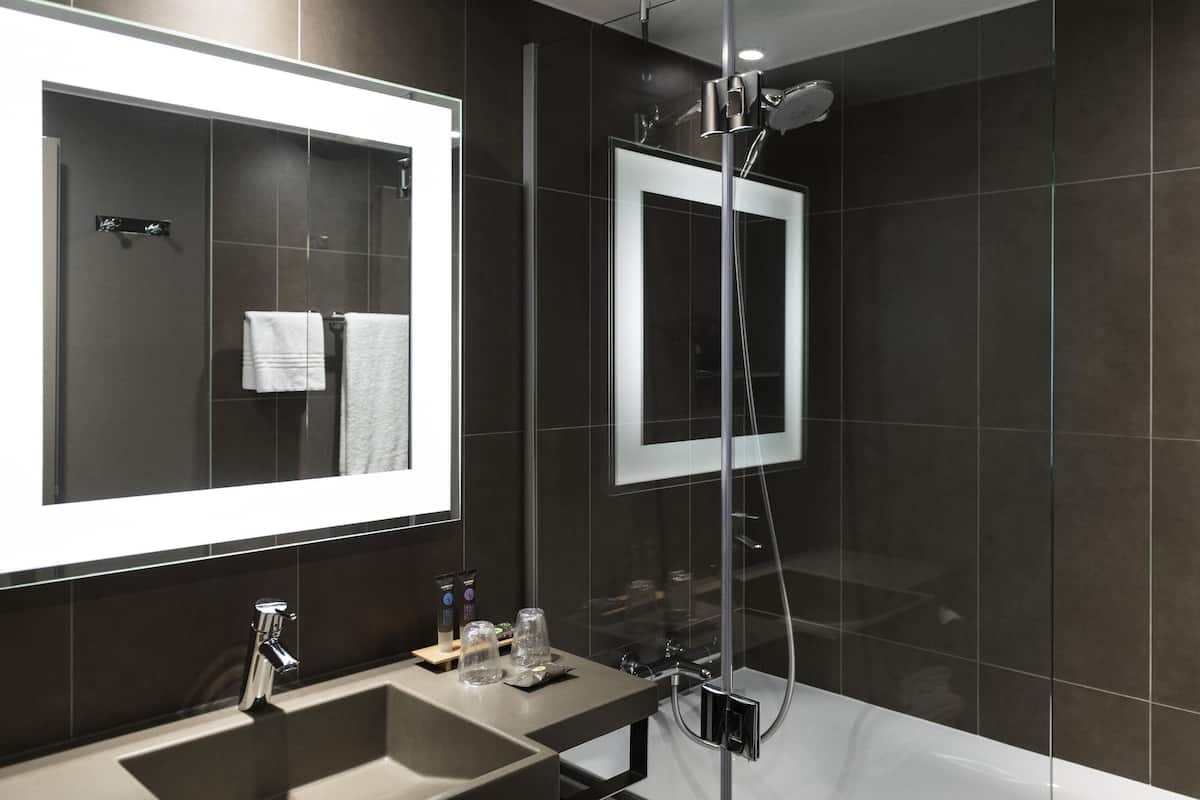 A modern bathroom with a large mirror, a sink, a shower, and dark tiled walls.