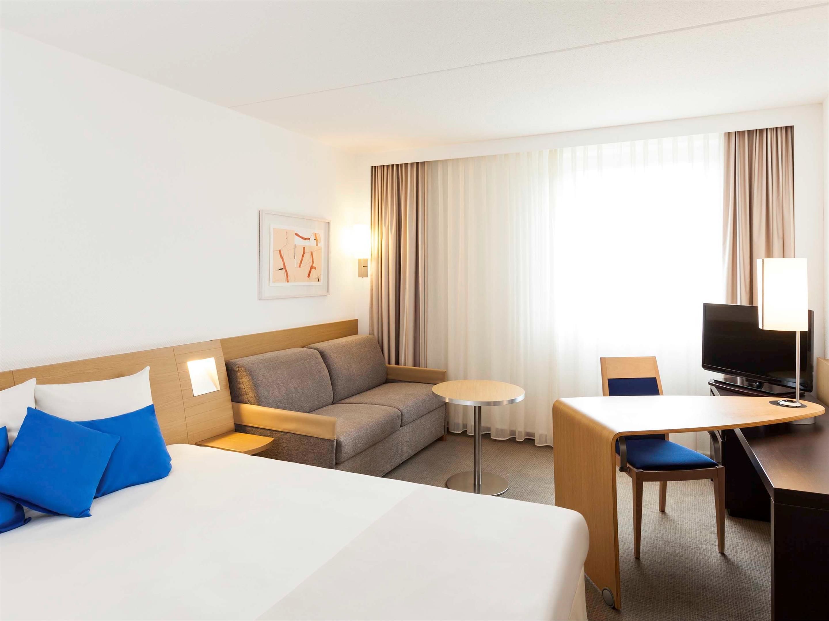 Habitación de hotel con cama, sofá, escritorio, silla, televisión y ventana con cortinas.