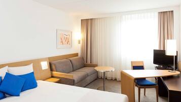 Habitación de hotel con cama, sofá, escritorio, silla, televisión y ventana con cortinas.