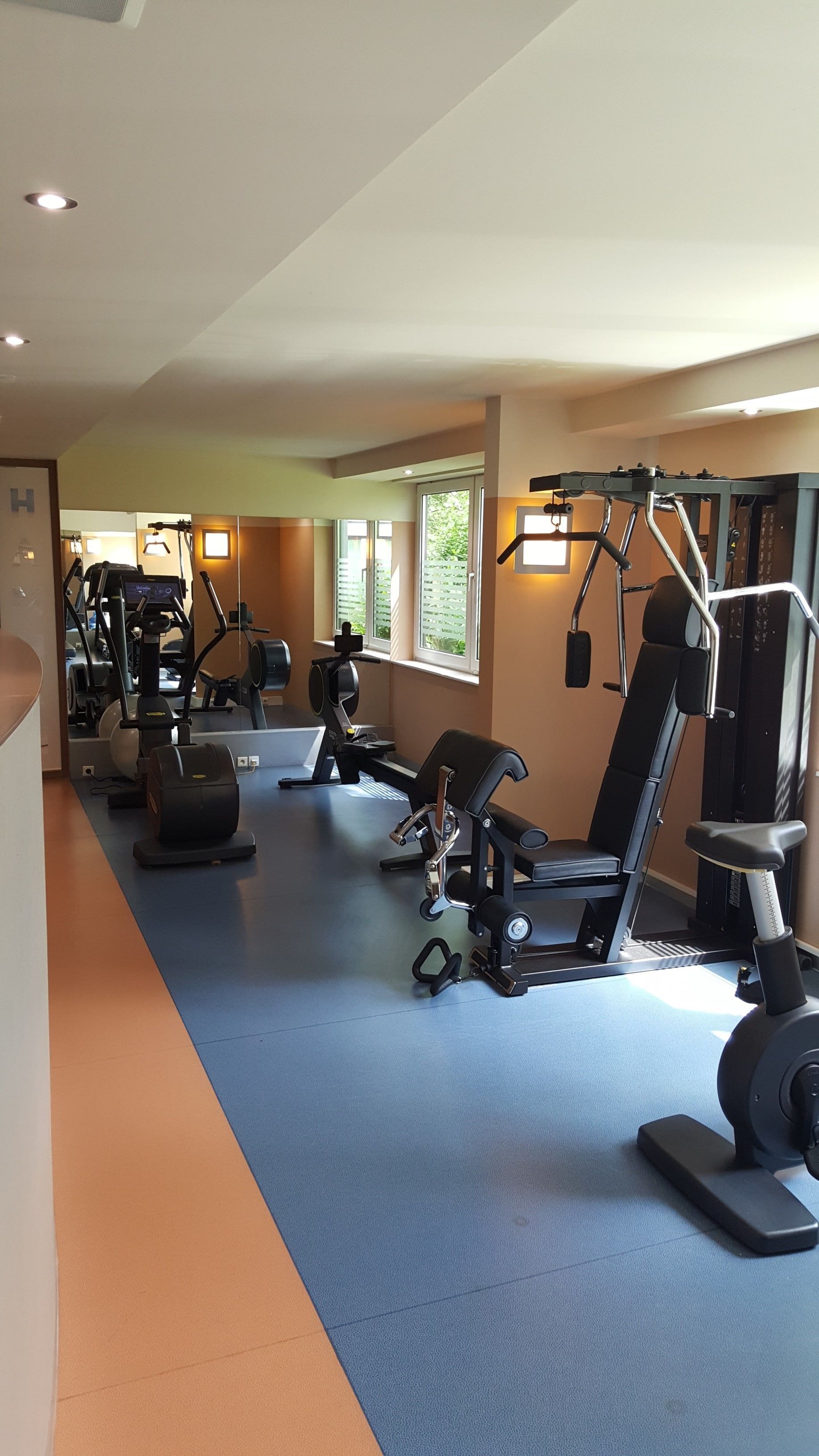 Une salle de sport équipée de divers appareils d’exercice, dotée d’un sol bleu et orange et de grandes fen êtres laissant entrer la lumière naturelle.