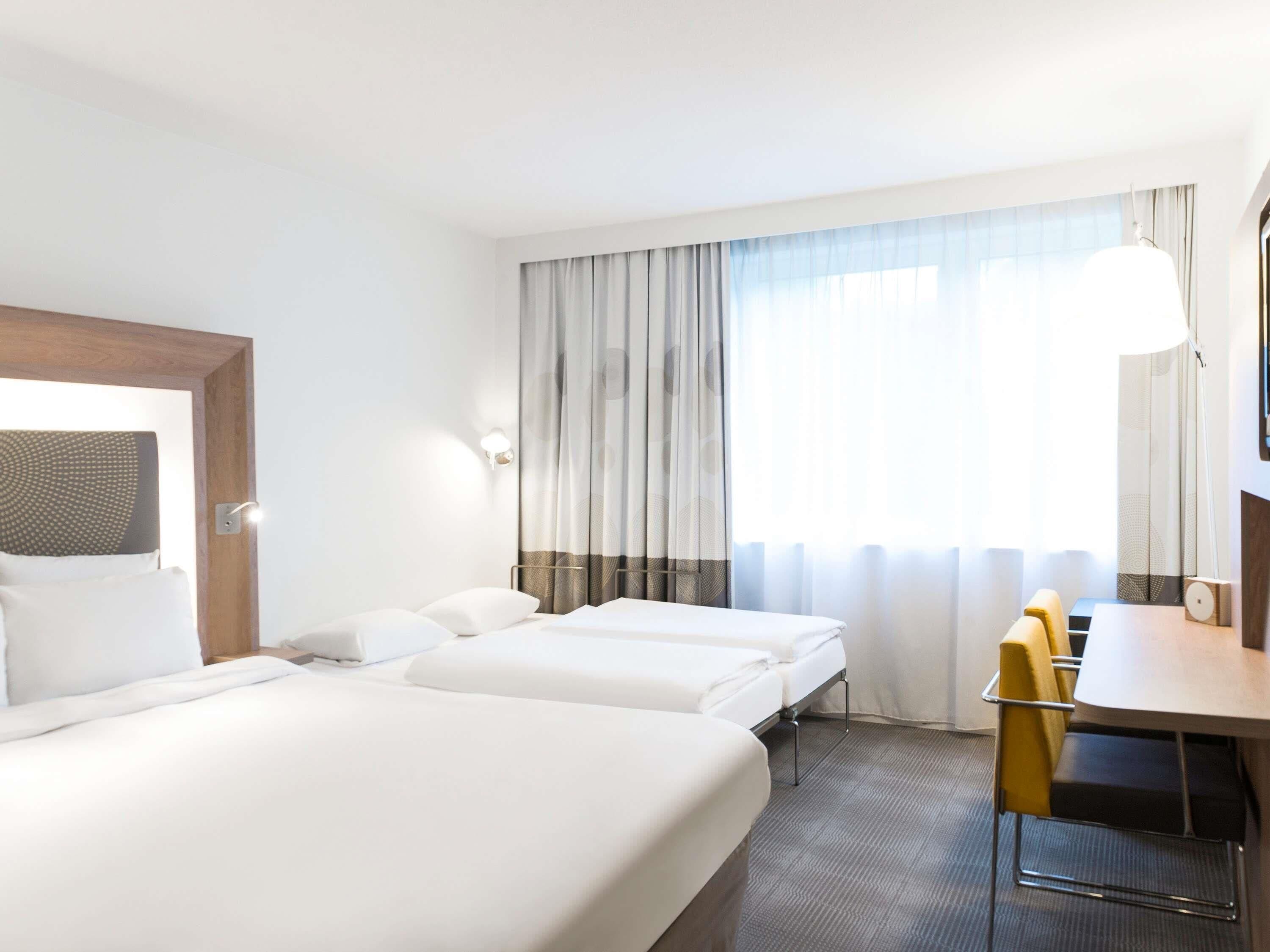 Ein Hotelzimmer mit zwei Betten, einem Schreibtisch, einem Sessel und einem großen Fenster mit Vorhängen.
