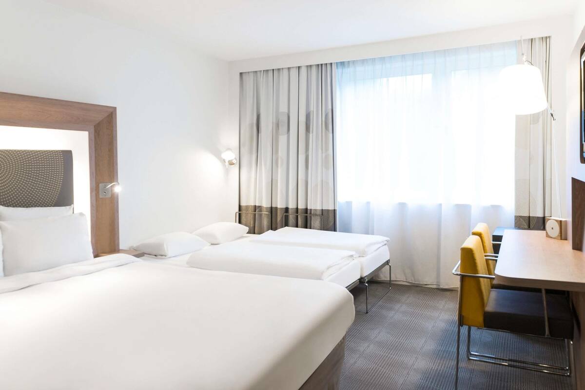 A hotel room with two beds, a desk, a chair, and a large window with curtains.