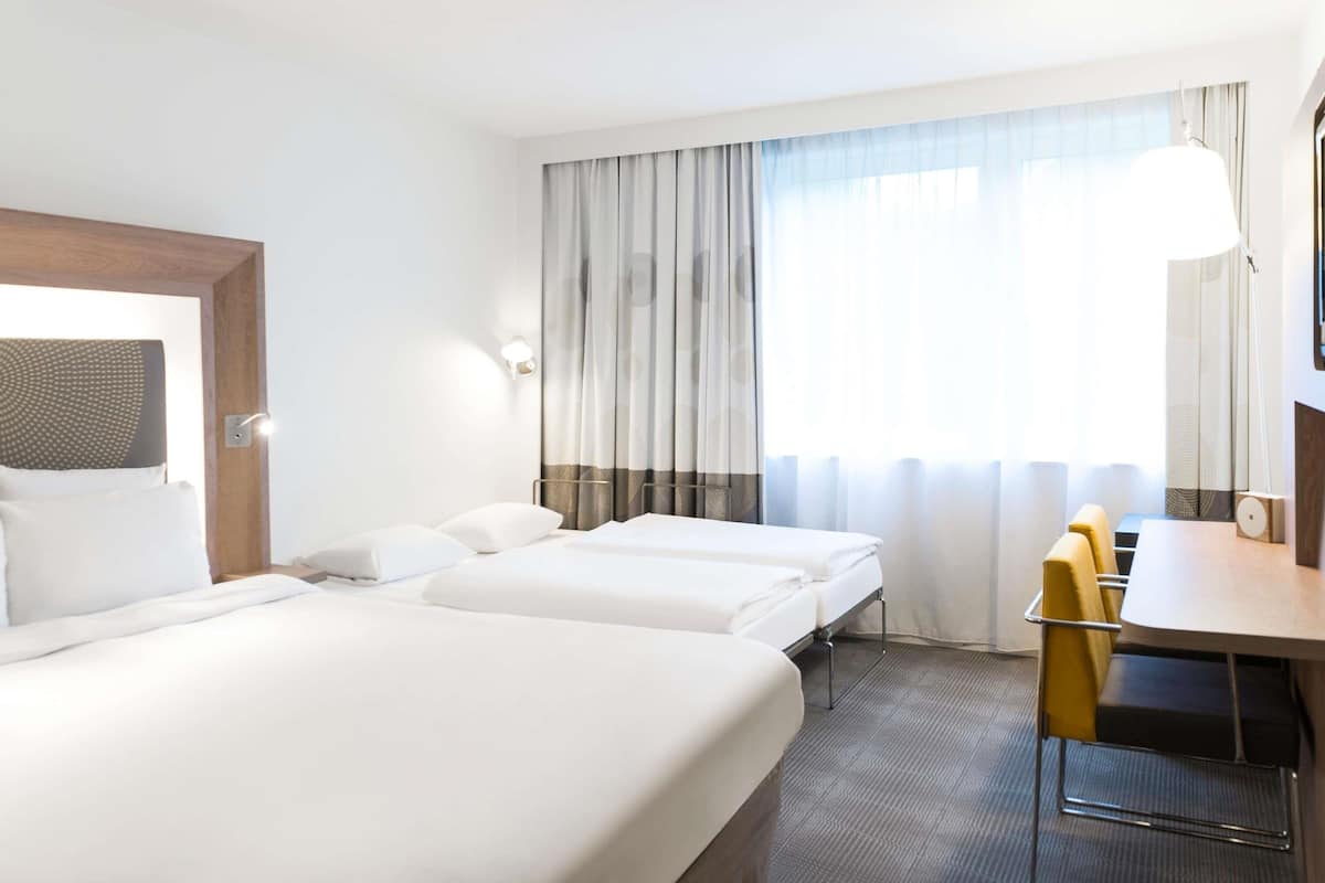 Ein Hotelzimmer mit zwei Betten, einem Schreibtisch, einem Sessel und einem großen Fenster mit Vorhängen.