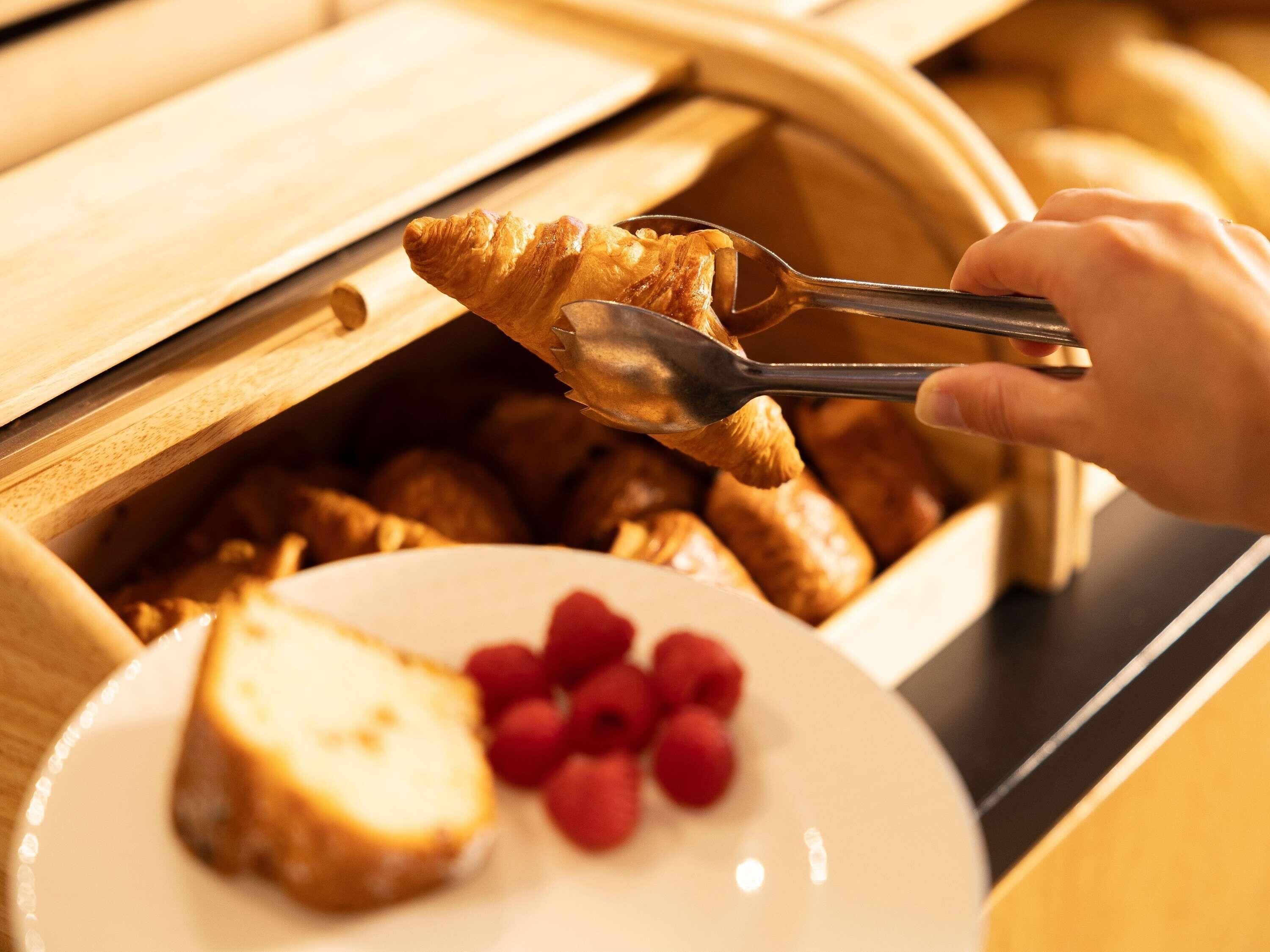 Une main utilisant des pinces pour servir de la nourriture contenue dans un récipient en bois, avec une assiette de nourriture et des framboises visibles.