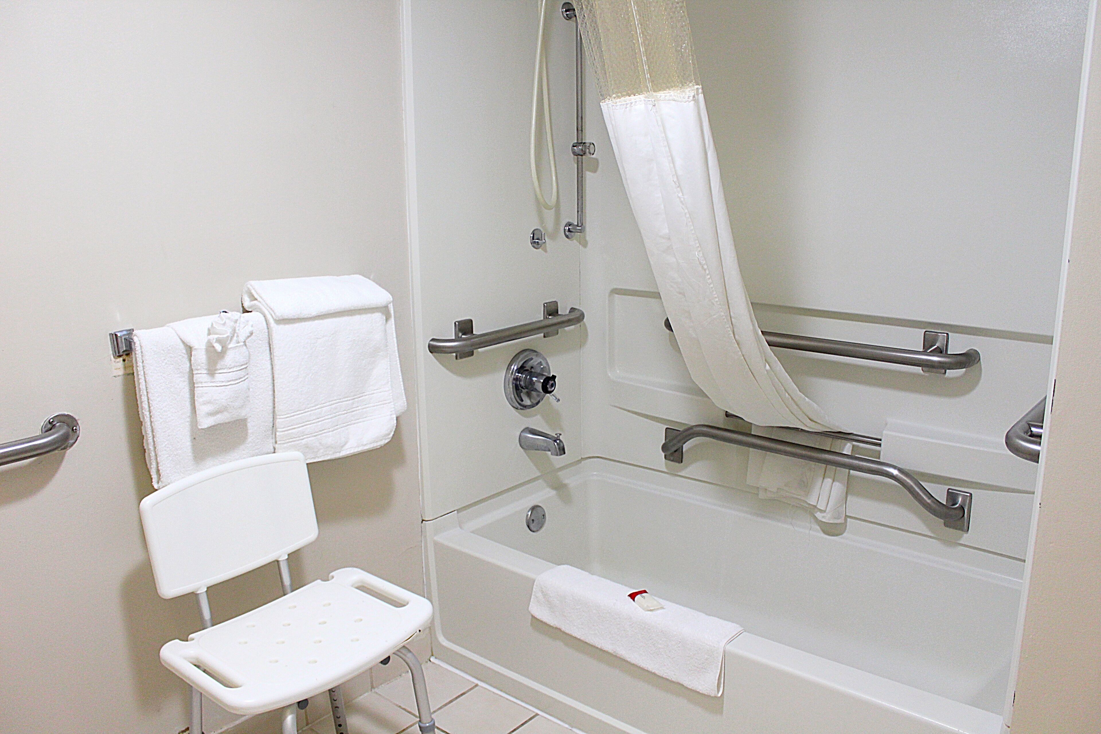 A bathroom with a bathtub, grab bars, a white chair, and a shower curtain.