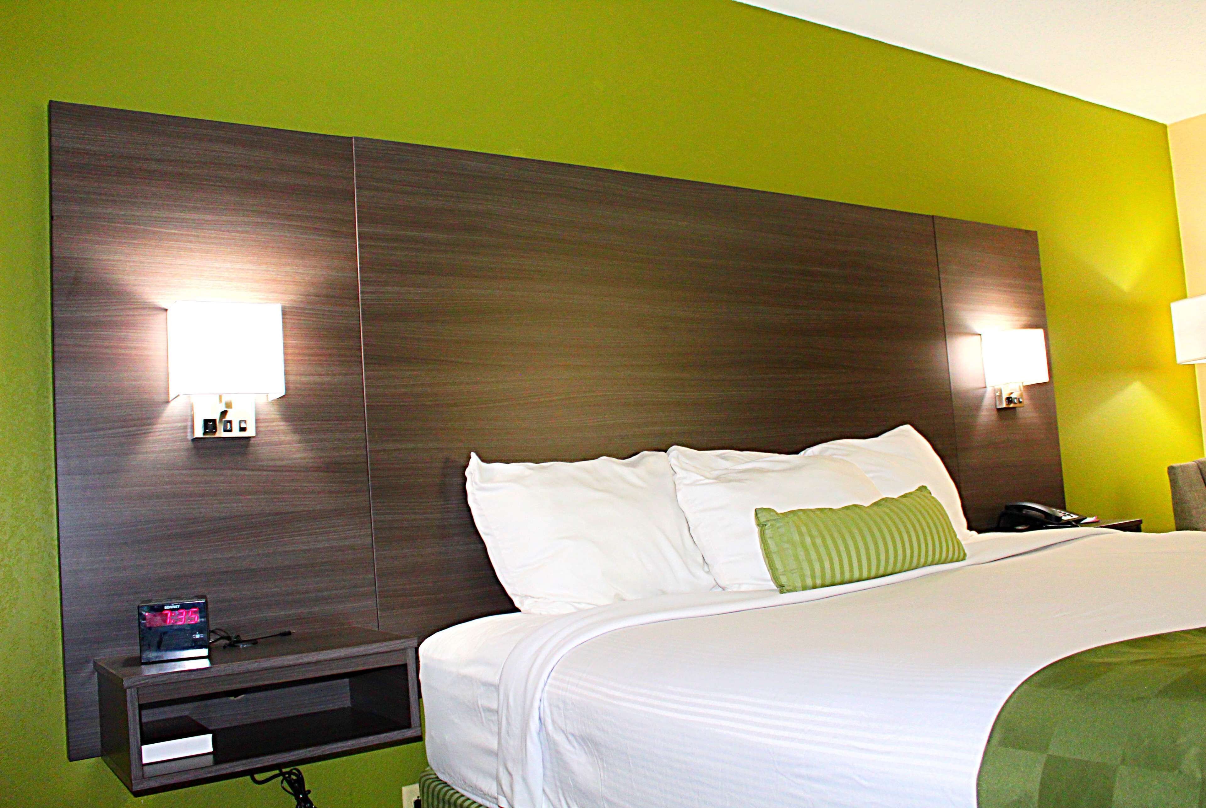Quarto de hotel com uma cama grande, duas luminárias de cabeceira, mesa de cabeceira com relógio e parede verde.
