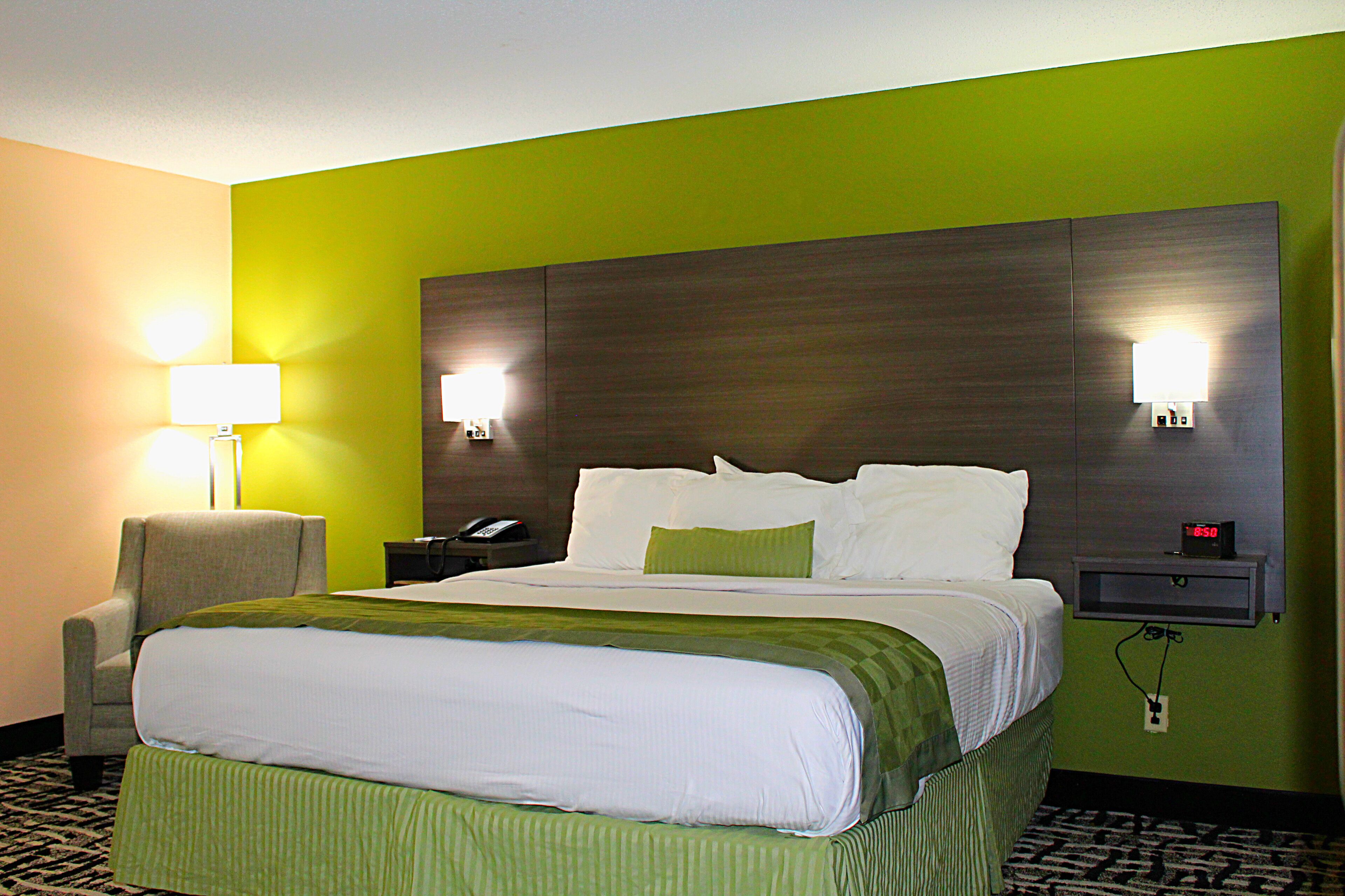 A hotel room with a large bed, green walls, two bedside lamps, a chair, a nightstand with a phone, and a clock.