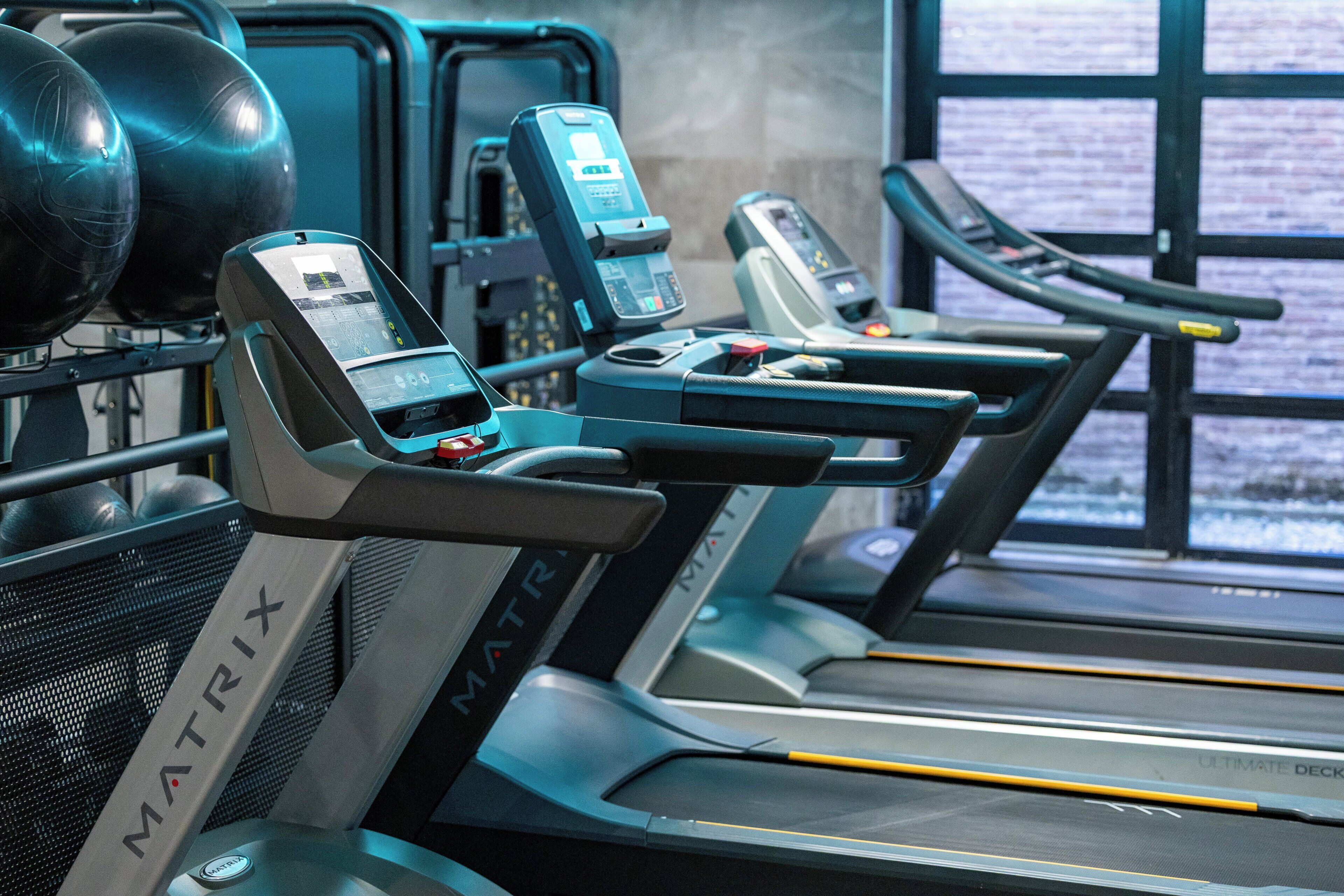 A row of treadmills with digital displays, a control panel, and a window with a view of the outside.