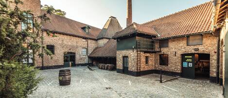 A cobblestone courtyard with brick buildings, a tall chimney, and a wooden barrel.
