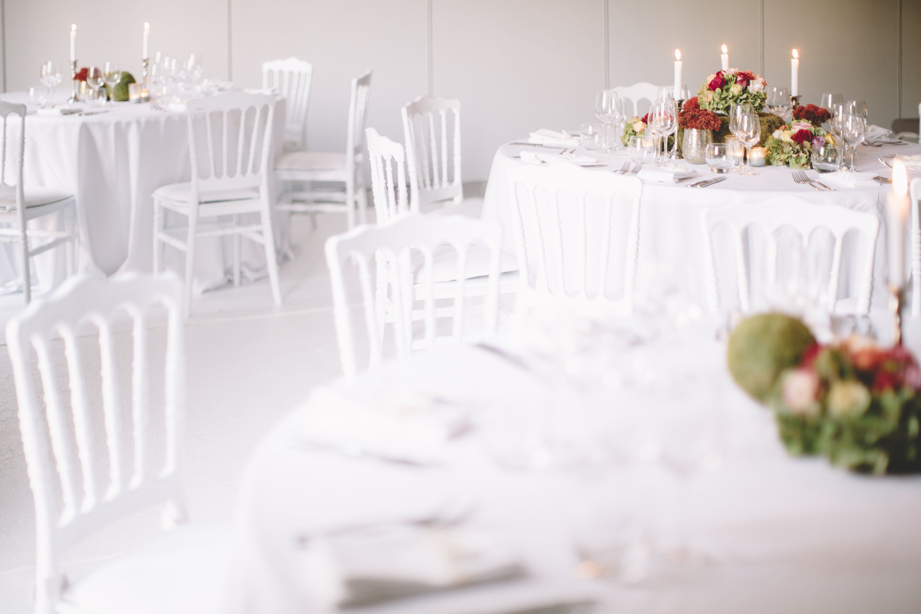 A room with round tables set for dining, white chairs, and a floral centerpiece.