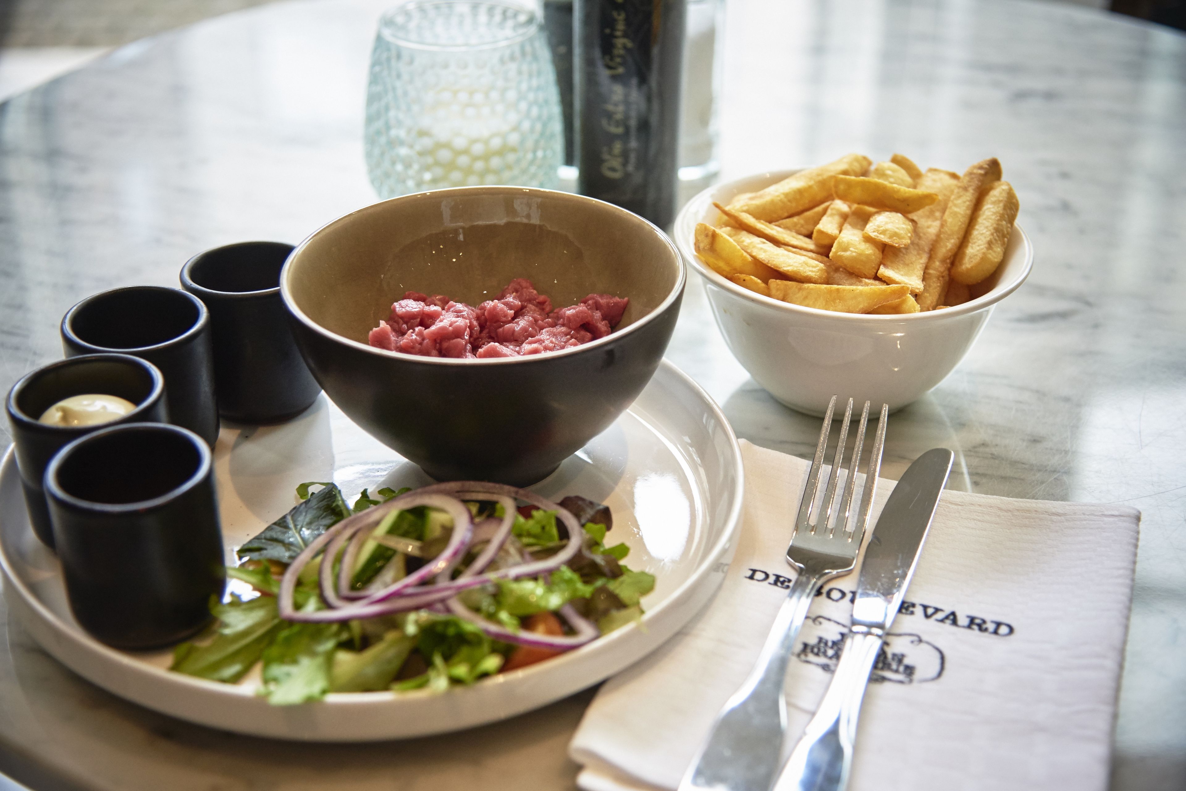 A table with a plate of salad, a bowl of diced meat, a bowl of French fries, and a napkin with a fork and knife.