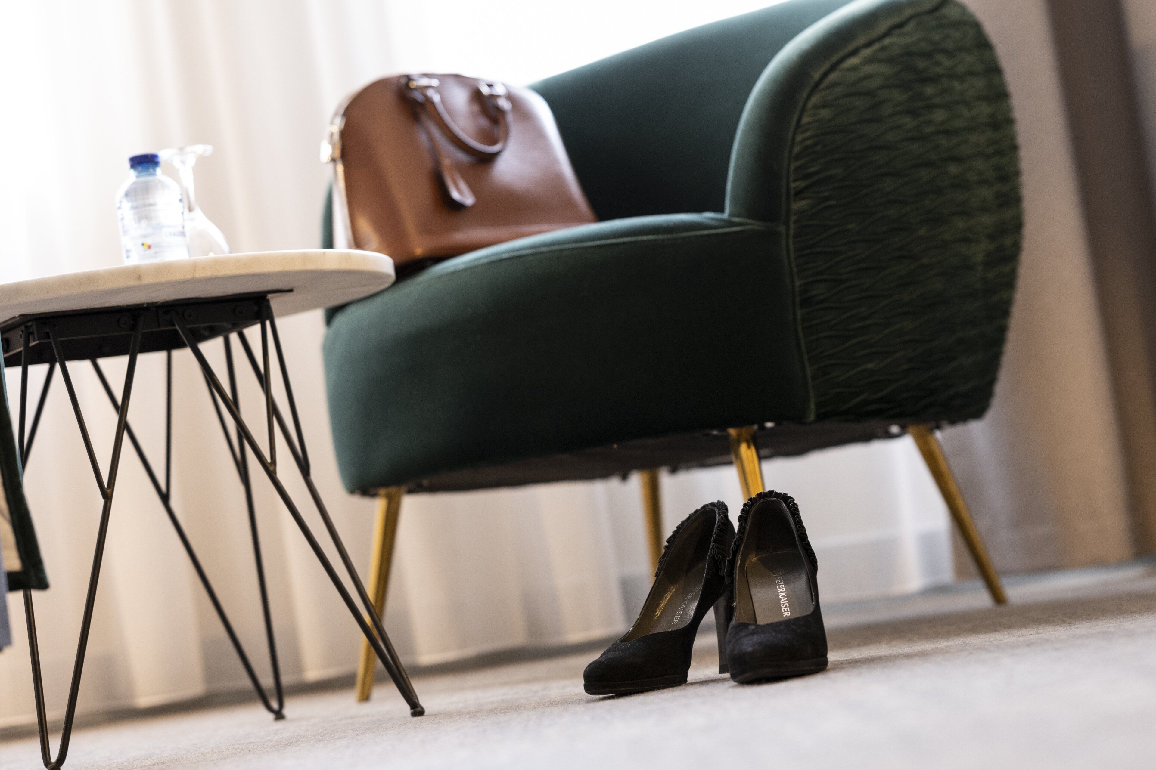 Un fauteuil vert avec un sac à main marron posé dessus, une paire d’escarpins noirs et une bouteille d’eau sur une petite table.