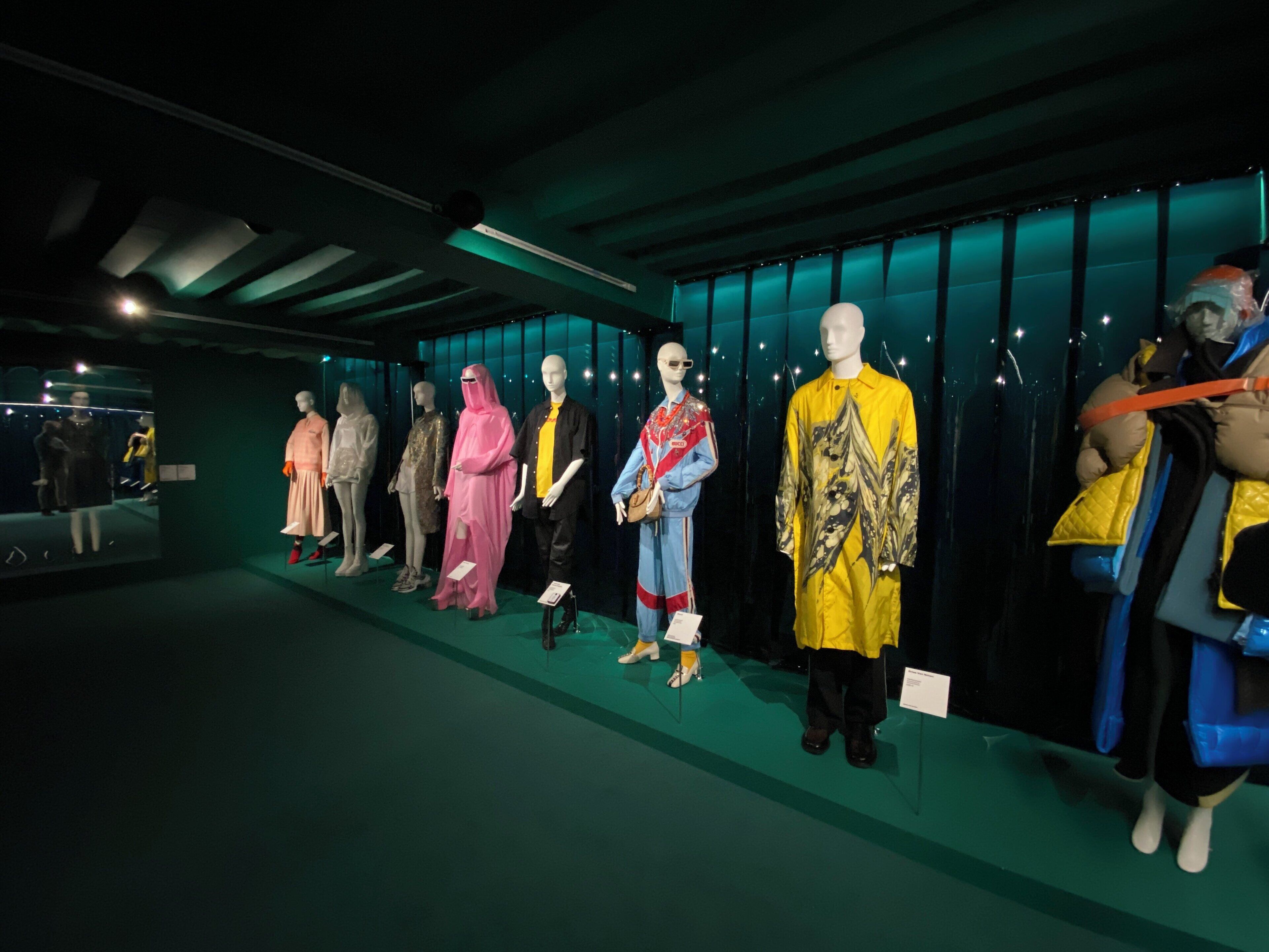 Poppen etaleren verschillende outfits in een schemerige ruimte met een groene vloer en een reflecterende muur.