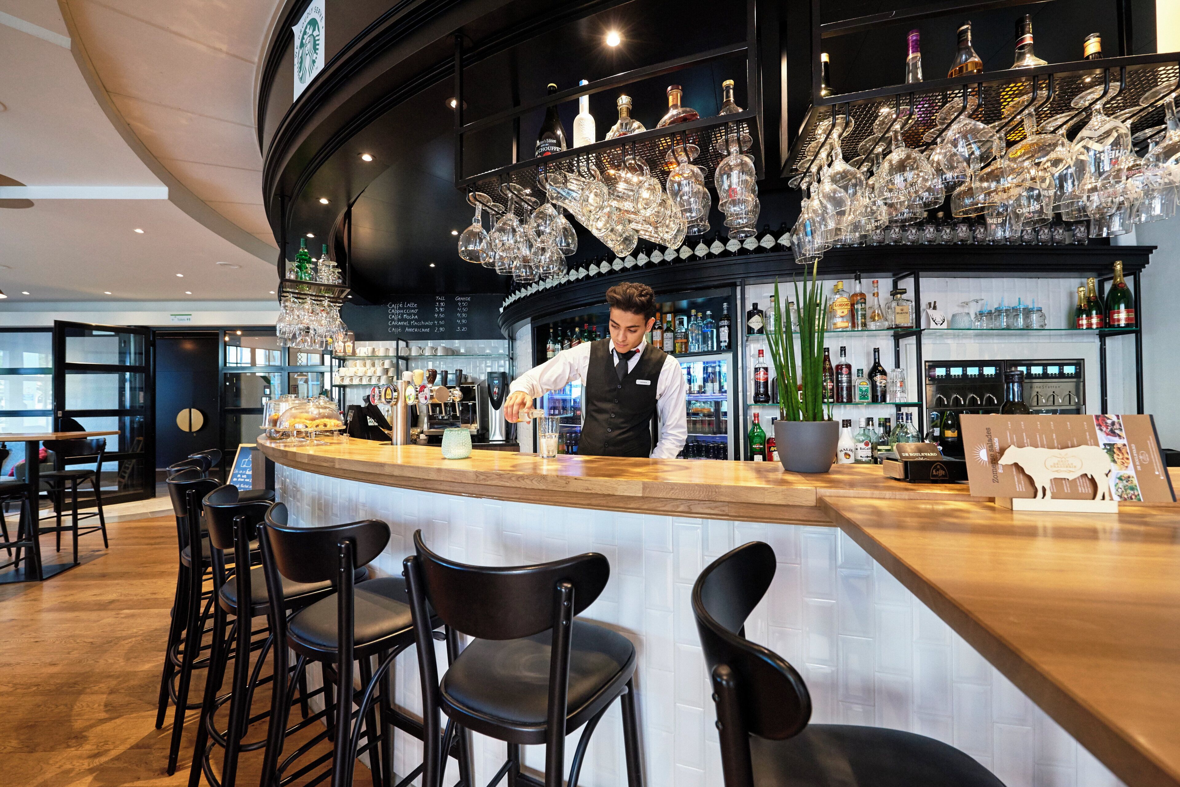 Zona de bar con un cantinero, taburetes, una variedad de botellas y una lámpara de cristal decorativa.