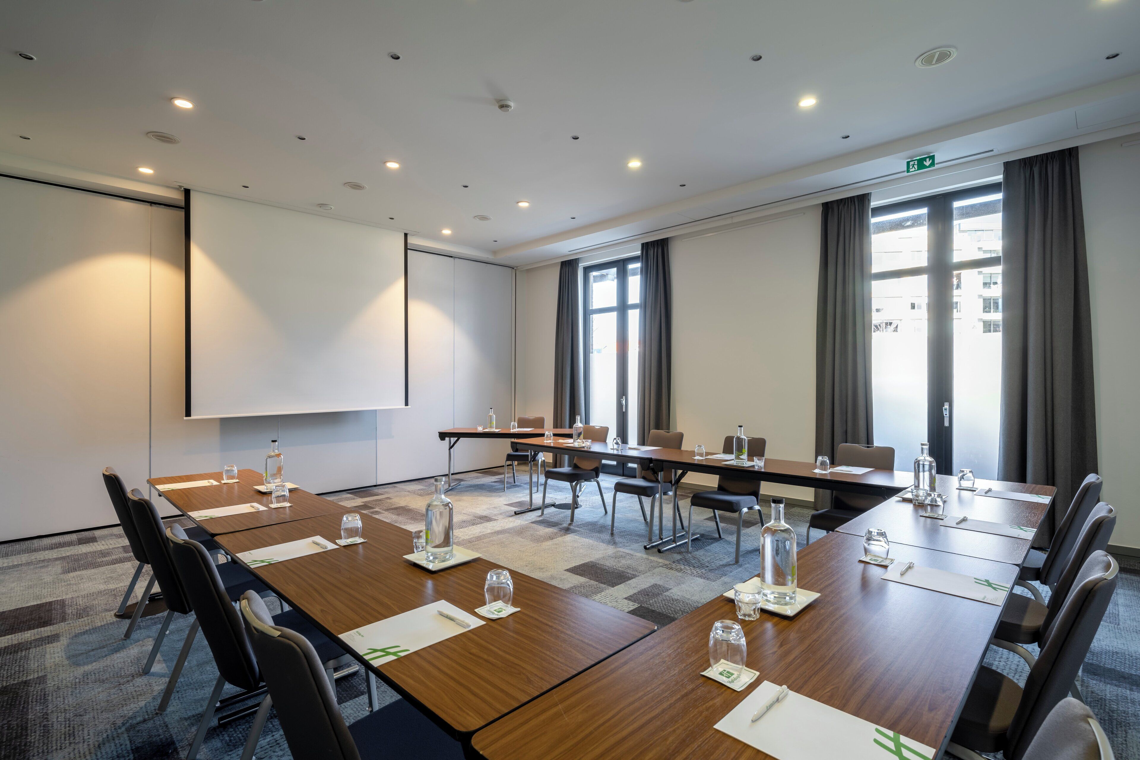 Una sala de conferencias con una larga mesa, sillas, una pantalla de proyector y un amplio ventanal con cortinas.