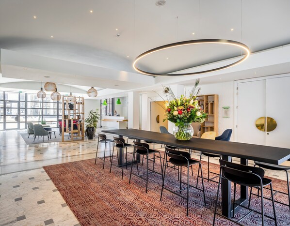 Een moderne hotel lobby met een lange eettafel, meerdere stoelen, een grote bloemenvaas en een receptiebalie.