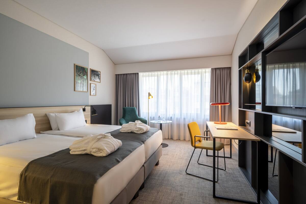 Een hotelkamer met een bed, een bureau, een stoel, een lamp en een raam met gordijnen.