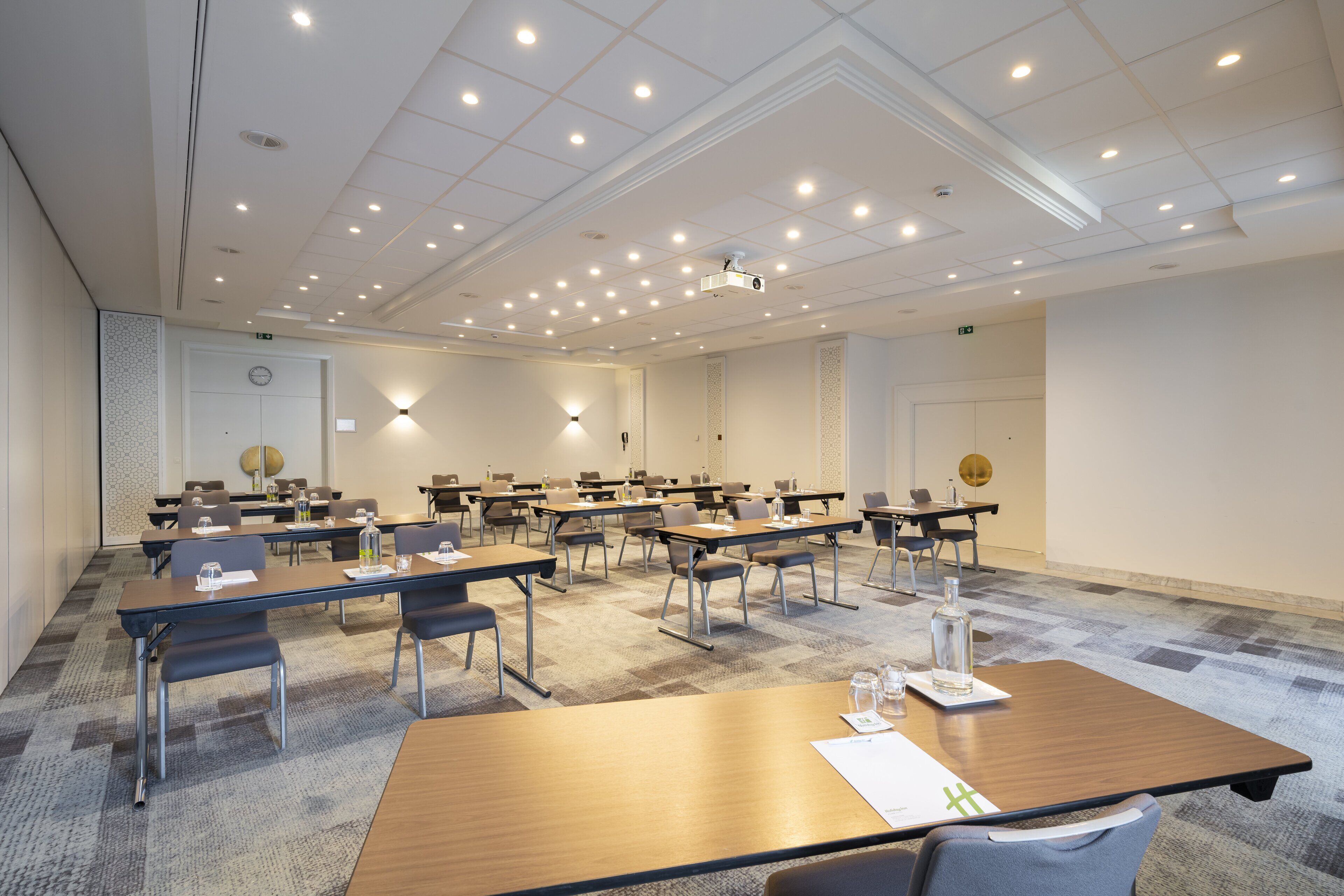 Une salle de conférence dotée de plusieurs tables et chaises, équipée de bouteilles d’eau et de documents, et un projecteur installé au plafond.