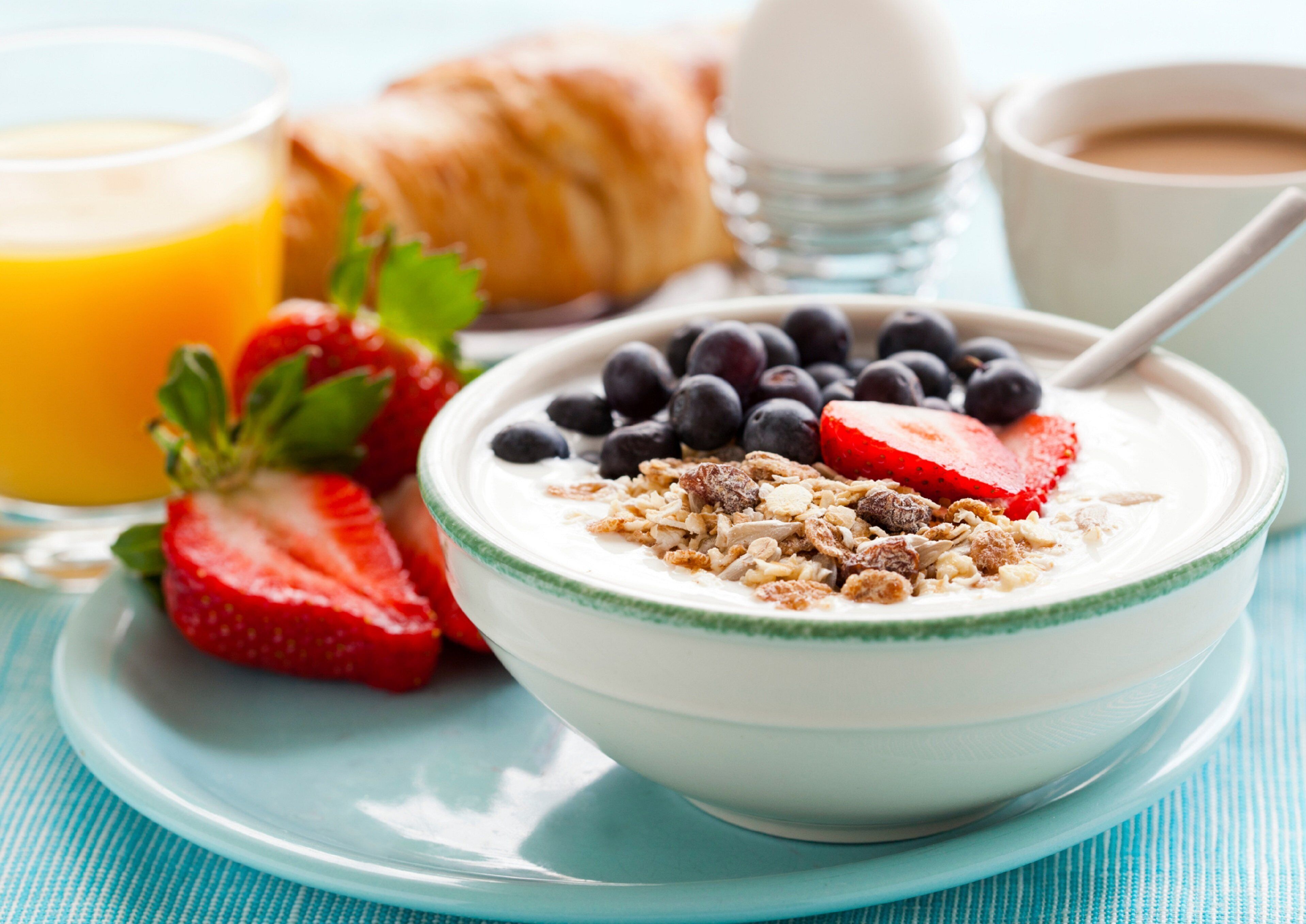 A bowl of yogurt with blueberries and strawberries, a croissant, a glass of orange juice, and a cup of coffee on a blue tablecloth.