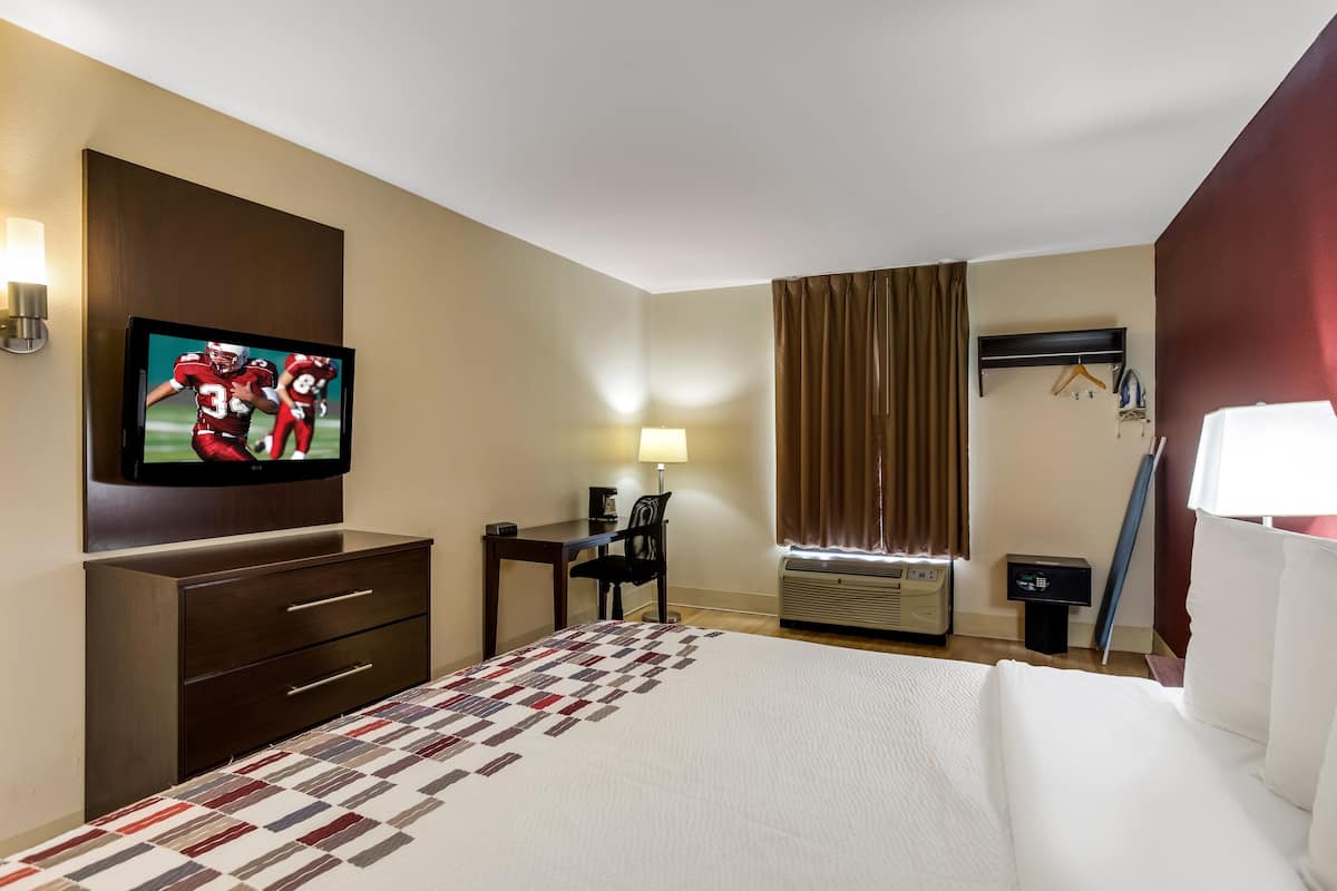 A hotel room with a large bed, a desk with a chair, a television mounted on the wall, and an air conditioning unit.