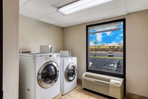 Une buanderie équipée de deux lave-linge professionnels de marque LG, d’une fenêtre donnant sur un parking et un bâtiment FedEx, ainsi que d’un climatiseur fixé au mur.