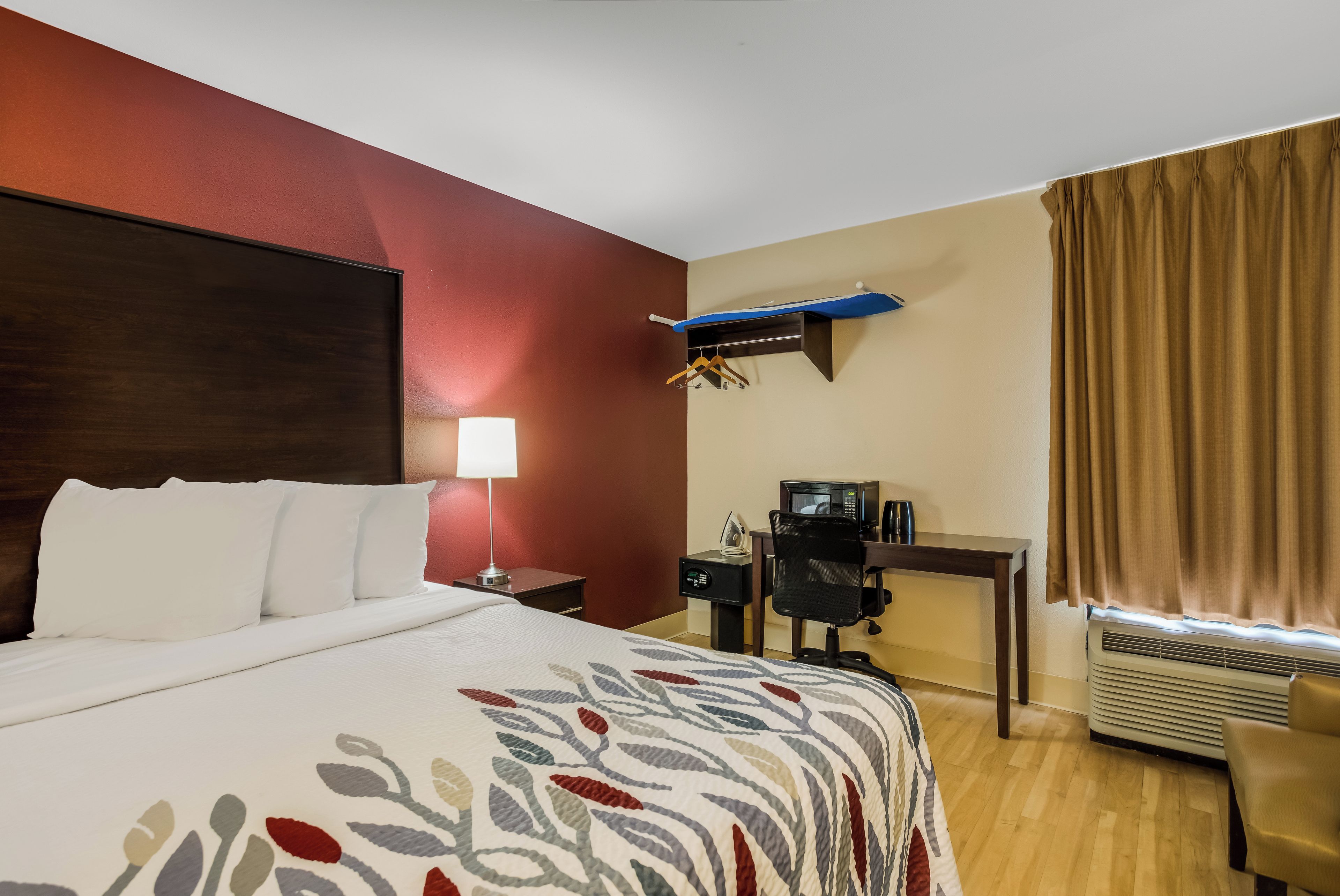 A hotel room with a bed, desk, chair, lamp, and air conditioning unit.