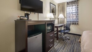 Habitación de hotel con televisión de pantalla plana, un mini refrigerador, un escritorio con silla, lámpara y ventana con cortinas estampadas.