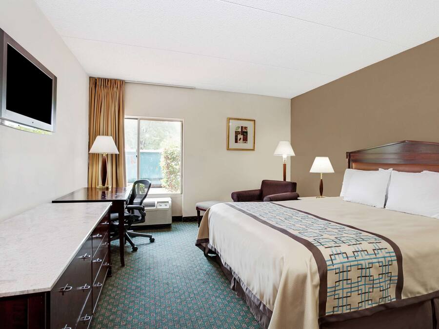 Ein Hotelzimmer mit einem großen Bett, einem Schreibtisch, zwei Stühlen, einem Fenster mit Vorhängen, einem gerahmten Bild an der Wand und einem an der Wand befestigten Flachbildfernseher.