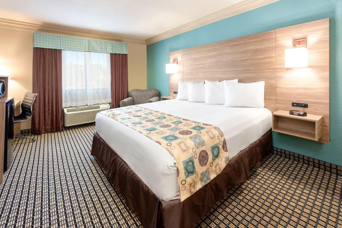 A hotel room with a large bed, a desk with a chair, a TV, a window with curtains, and a wall-mounted shelf.