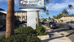Señal que dice 'PB SURF BEACHSIDE INN', con un gráfico de tabla de surf, rodeada de palmeras y un cielo despejado.