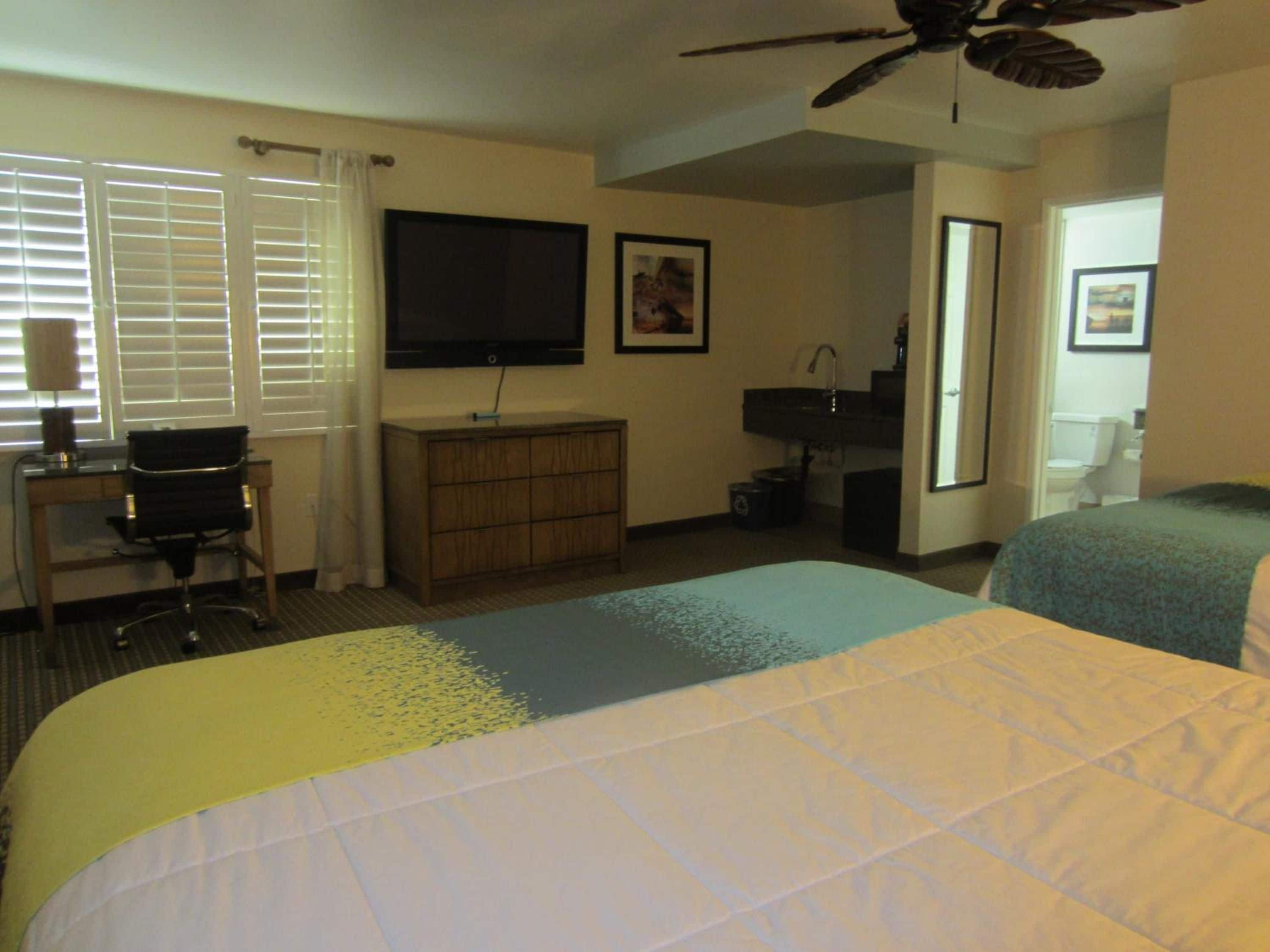 A hotel room with two beds, a desk with a chair, a television, a ceiling fan, and a bathroom.