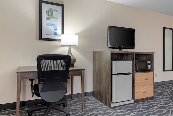 A hotel room with a desk, chair, TV, refrigerator, microwave, and a framed picture on the wall.