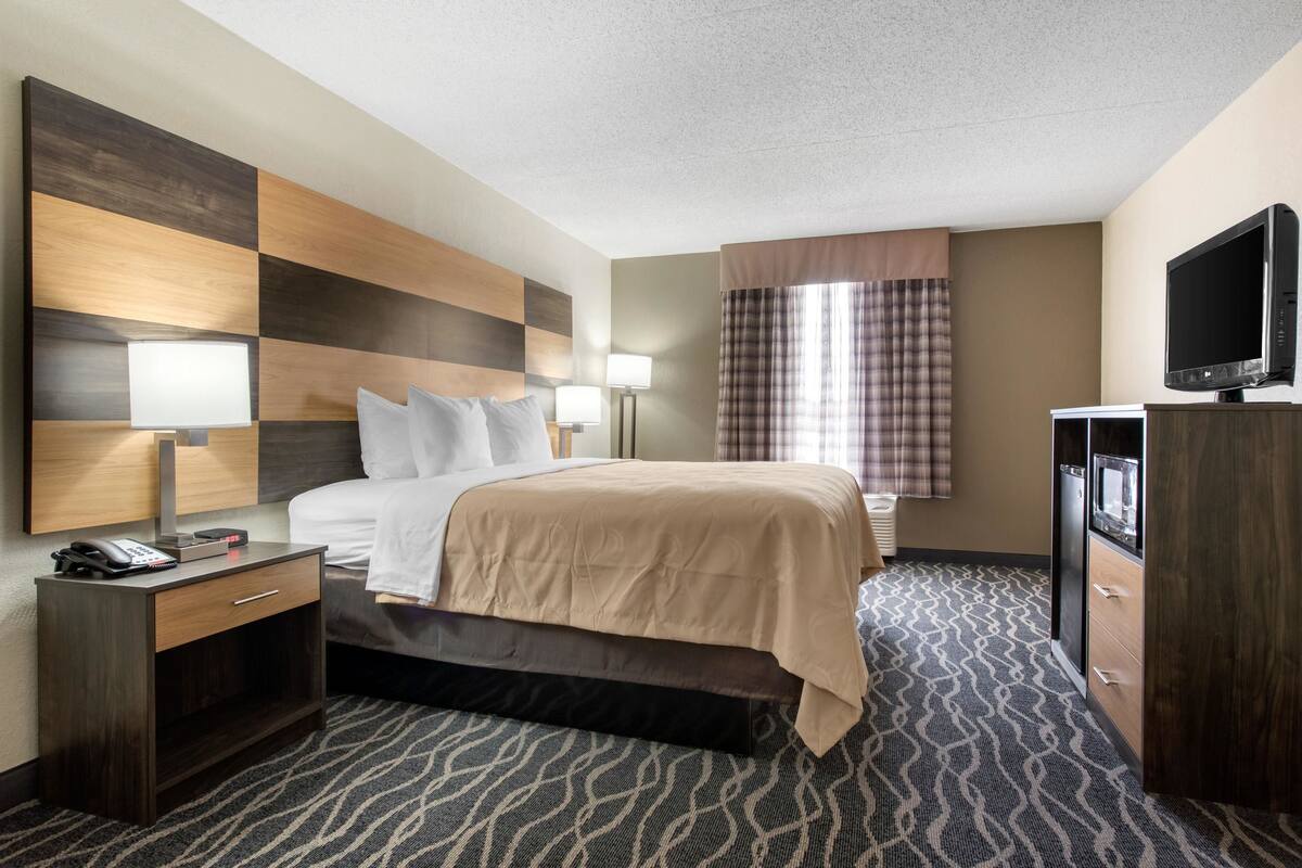 A hotel room with a bed, bedside lamps, a TV, a desk with a phone, and a patterned carpet.