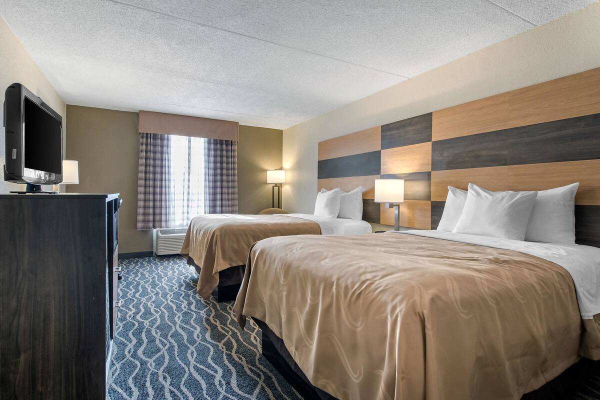A hotel room with two beds, a TV, a desk, and a window with curtains.
