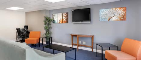 A modern office space with a grey couch, orange chairs, a black coffee table, and a TV on the wall.