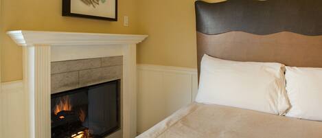 A bedroom with a fireplace, a bed with white bedding, and a wall-mounted artwork.