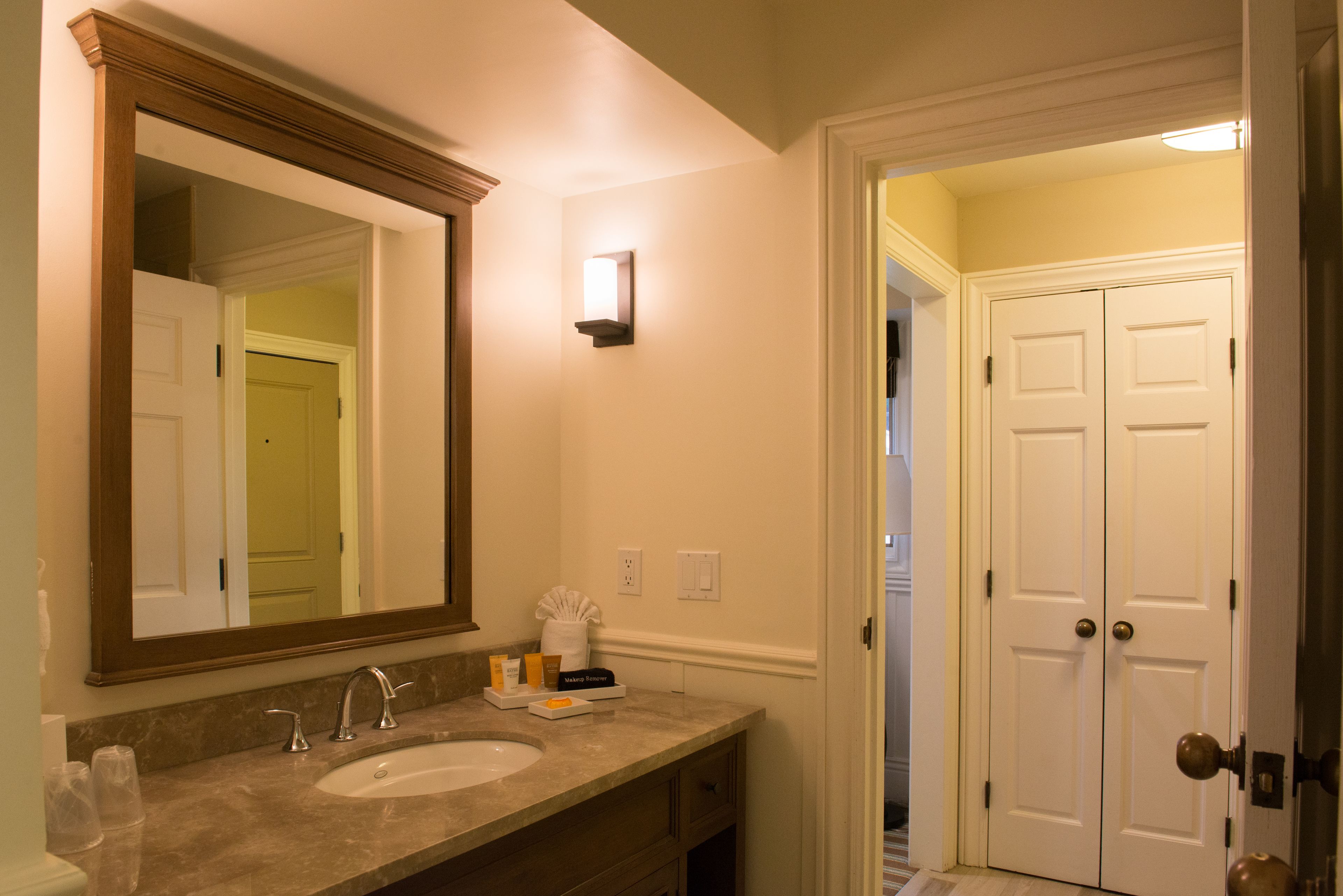 Une salle de bain avec un grand miroir, un lavabo et un porte-serviettes. Une porte donne accès à une autre pièce.