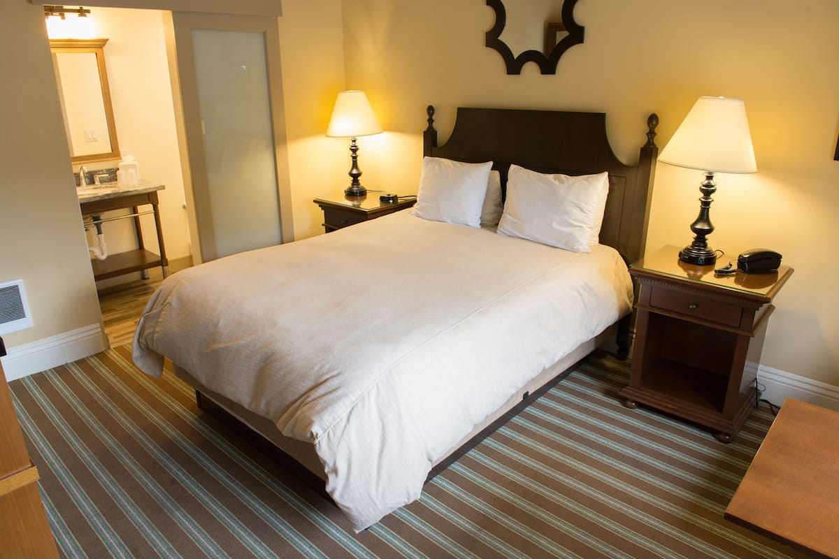 A neatly made bed with white linens, two bedside tables with lamps, a wooden headboard, and a striped carpet.