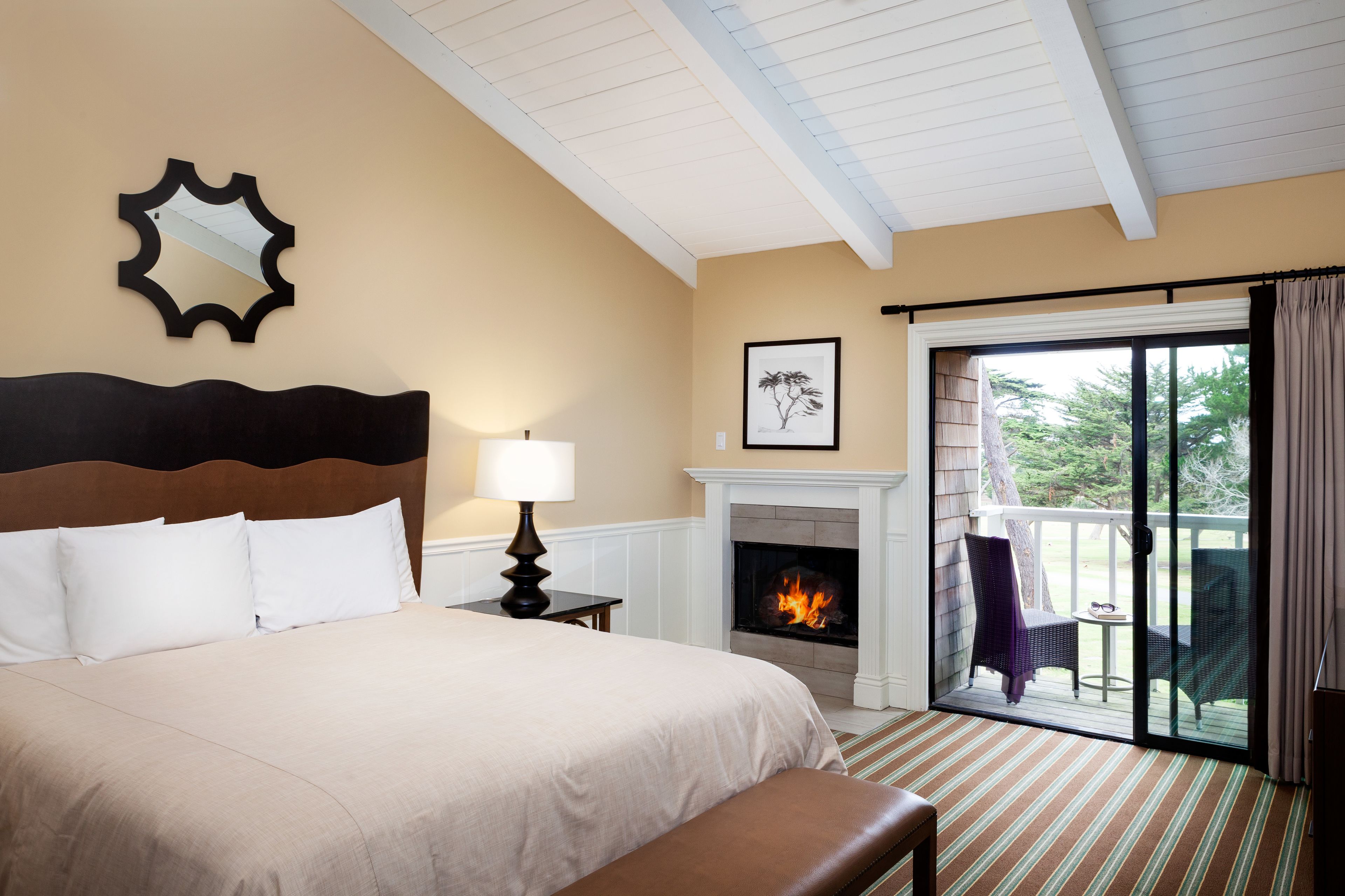 A hotel room with a bed, a fireplace, a balcony with chairs, and a view of the outdoors.