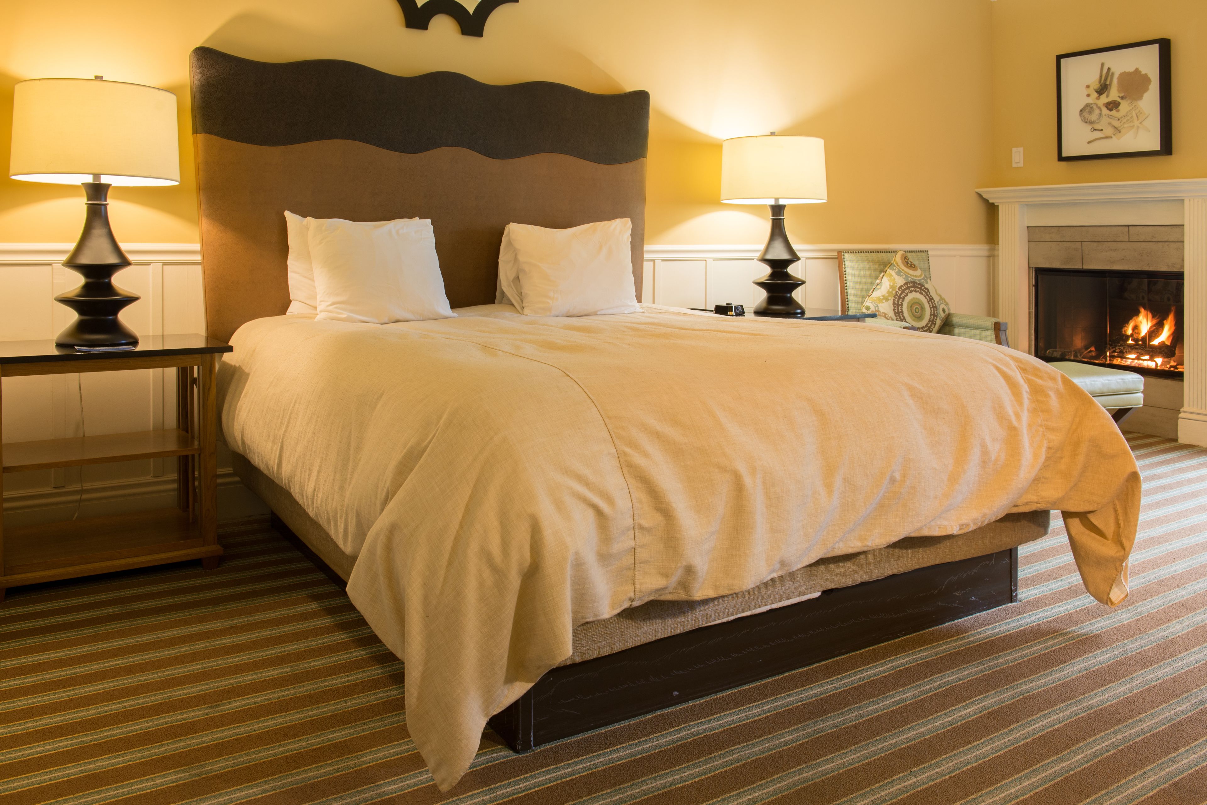A hotel room with a large bed, two bedside tables with lamps, a fireplace, and a striped carpet.