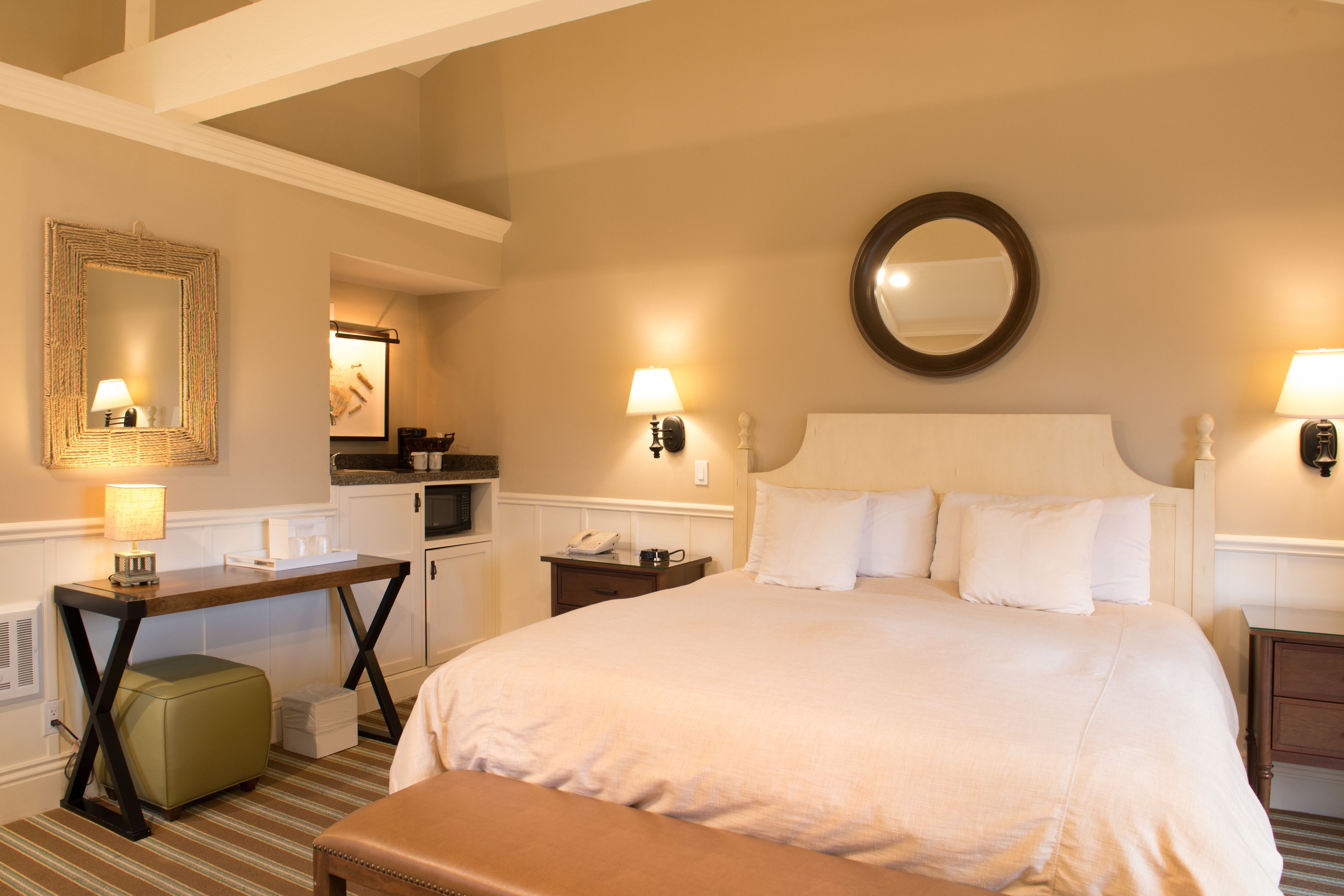 Une chambre d’hôtel avec un grand lit, un bureau, une chaise, un banc, un miroir et une petite kitchenette.